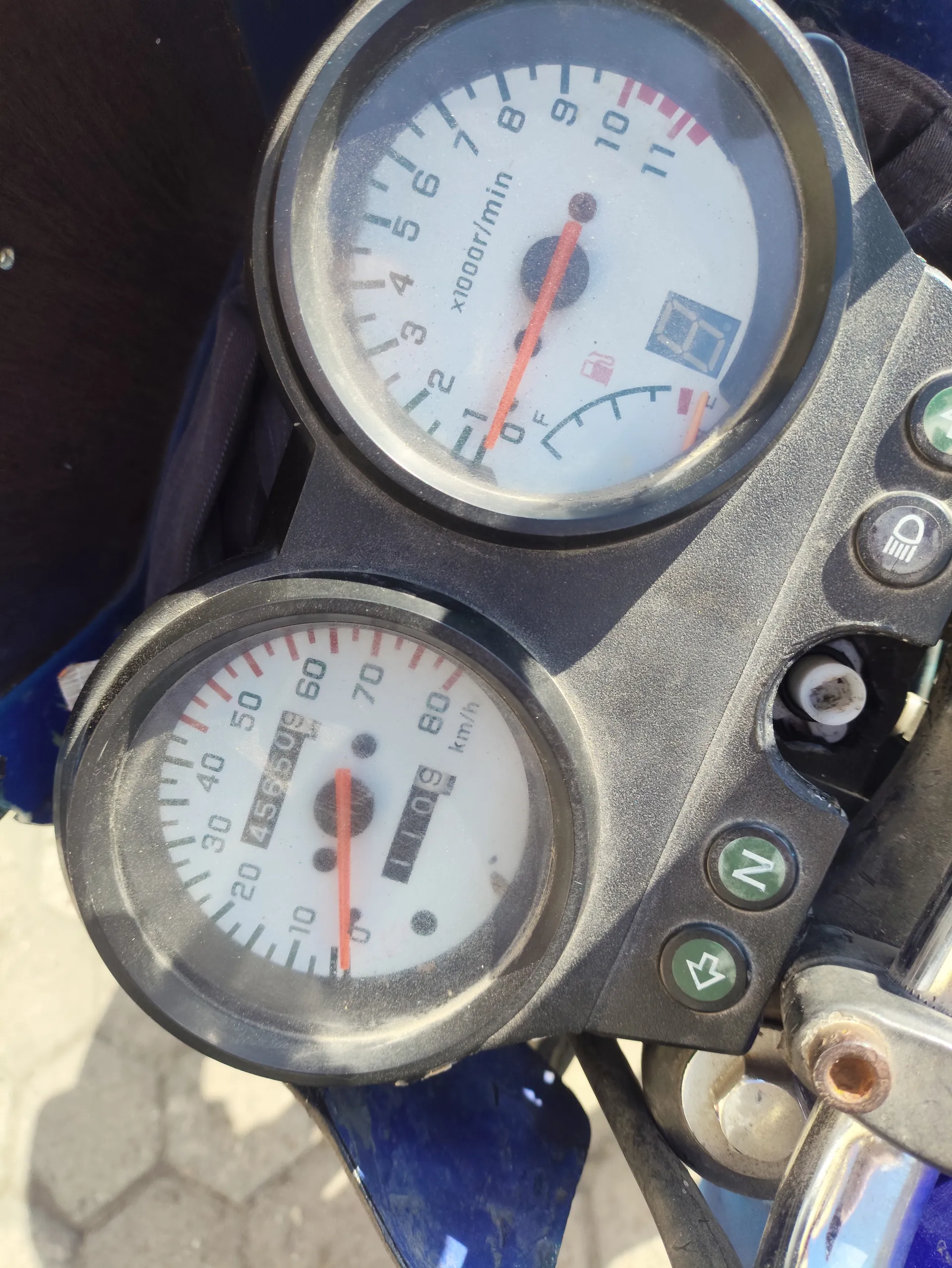 Zbliżenie na deski rozdzielcze motocykla. Górnym licznikiem jest tachometr z zakresem od 0 do 11 (oznaczony jako x1000r/min), z czerwoną igłą wskazującą blisko 0. Obok znajduje się cyfrowy wyświetlacz z wartością 0 i ikoną paliwa. Dolny licznik to prędkościomierz w km/h (zakres 0-80), z czerwoną igłą na 0, licznikiem przebiegu 45650,9 km i licznikiem dziennym 110,9 km. Po prawej stronie widoczne są przyciski: z literą N, zielonym strzałką, ikoną reflektora i innym zielonym przyciskiem. W tle fragment niebieskiego elementu motocykla i brukowanej nawierzchni.