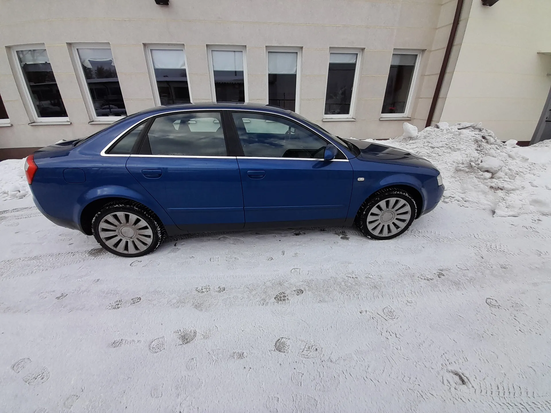 Niebieski sedan czterodrzwiowy zaparkowany na śnieżnej nawierzchni przed budynkiem o jasnej elewacji z szeregiem okien o białych ramach. Widoczne odciski stóp, ślady opon i czyste szyby. Po prawej wysoki wał śniegu. Samochód ma srebrne felgi i czarne listwy boczne.