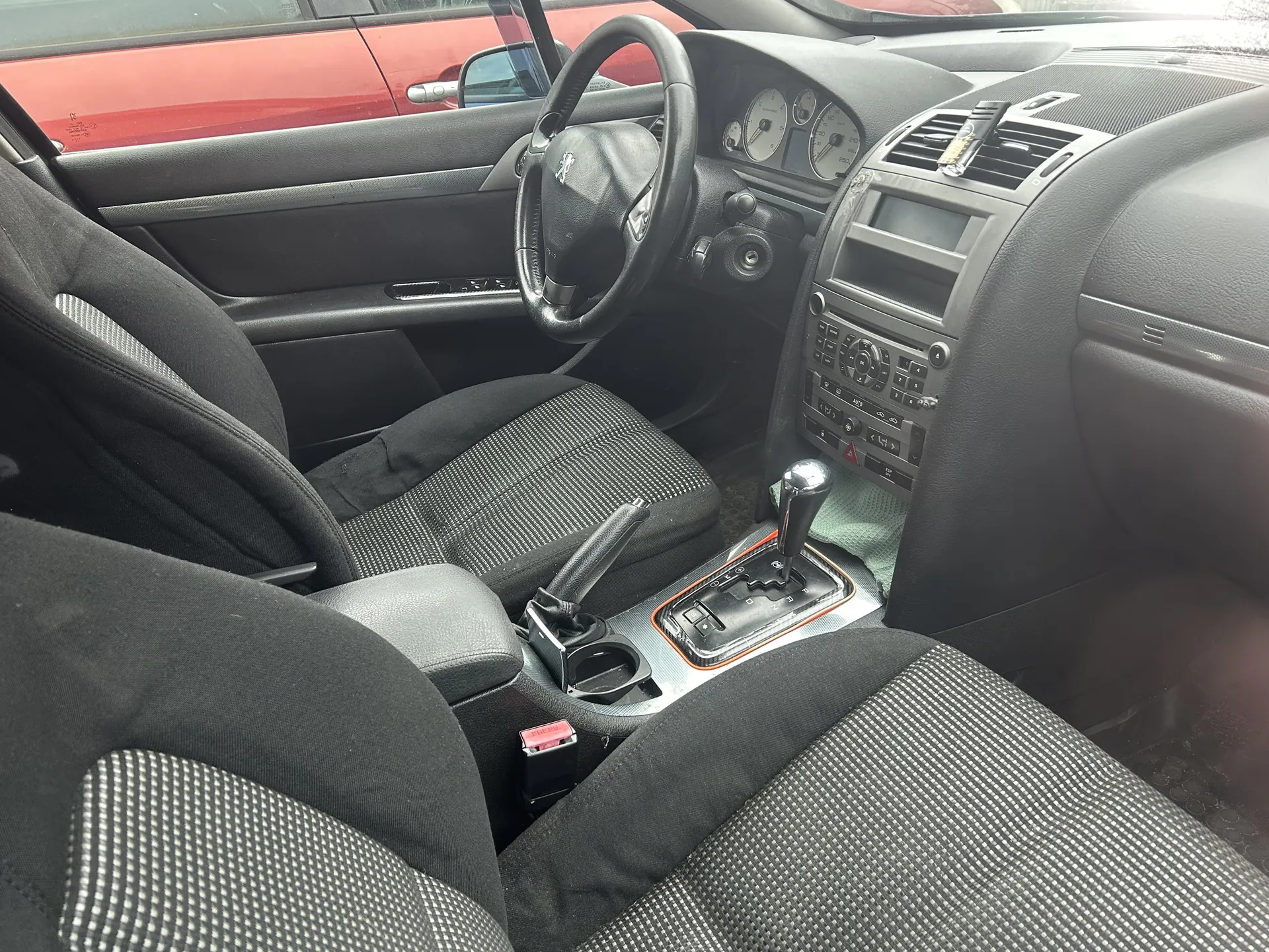 Wnętrze samochodu z czarnymi siedzeniami w szachownicowy wzór, kierownicą z logo, deską rozdzielczą z licznikami, centralną konsolą z przyciskami, dźwignią zmiany biegów i hamulcem ręcznym. Widoczne są również zewnętrzne elementy: czerwony samochód za oknem oraz fragmenty drzwi i deski rozdzielczej.