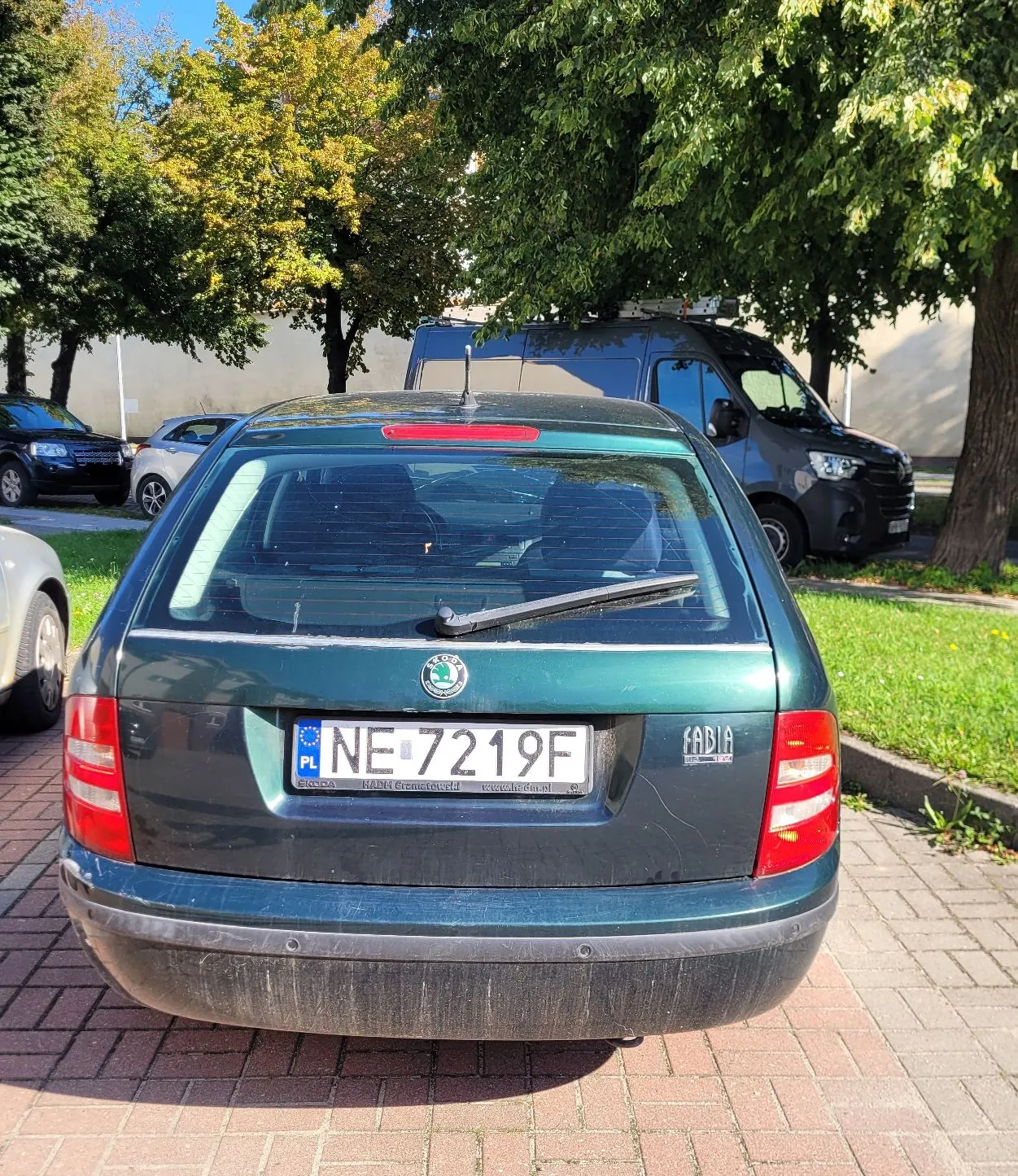 Tylny widok ciemnozielonego samochodu marki Skoda Fabia zaparkowanego na kostce brukowej. Samochód ma polską tablicę rejestracyjną o numerze NE 7219F. W tle widoczny jest srebrny samochód osobowy, szary samochód dostawczy oraz zielone drzewa.
