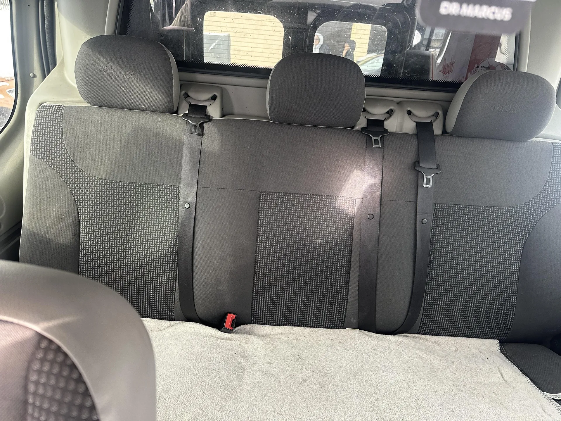 Wnętrze samochodu z widokiem na tylną kanapę. Trzy czarne zagłówki, pasy bezpieczeństwa z czarnymi paskami i srebrnymi zapięciami. Tapicerka siedzenia w czarno-szarym kolorze z drobnym wzorem. Na dole widoczna jasna tkanina. Przez tylne okno widać fragment budynku z drewnianą elewacją oraz ludzi. W prawym górnym rogu naklejka z napisem "GO MARCUS". W lewym dolnym rogu fragment przedniego siedzenia.