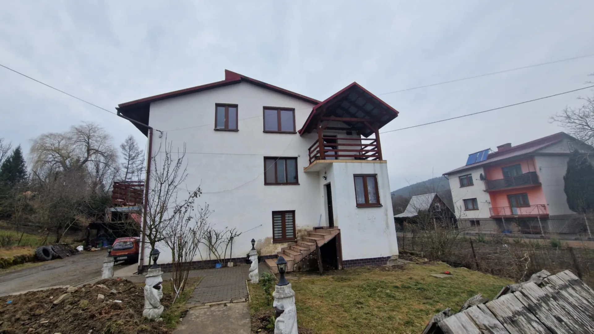 Biały dom dwupiętrowy z czerwonym dachem i drewnianym balkonem na prawej stronie. Dom posiada brązowe okna, schody prowadzące do wejścia oraz ścieżkę z kamiennymi filarami z latarniami. Przed domem rośnie bezlistne drzewo, po lewej stronie widoczny pomarańczowy samochód, opony i garaż. W tle domy mieszkalne, w tym jeden z niebieskim panelem słonecznym, drzewa i wzgórza. Niebo jest zachmurzone.