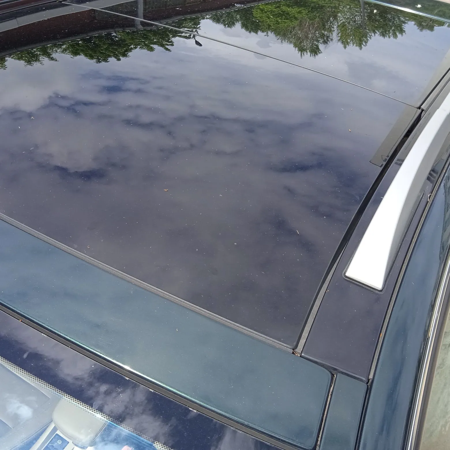 Zbliżenie na dach samochodu w ciemnozielonym kolorze z dużym, ciemnym szklanym panelem panoramicznym. W gładkiej powierzchni szkła odbija się niebo z chmurami oraz korony drzew. Po prawej stronie widoczna jest srebrna reling dachowa.