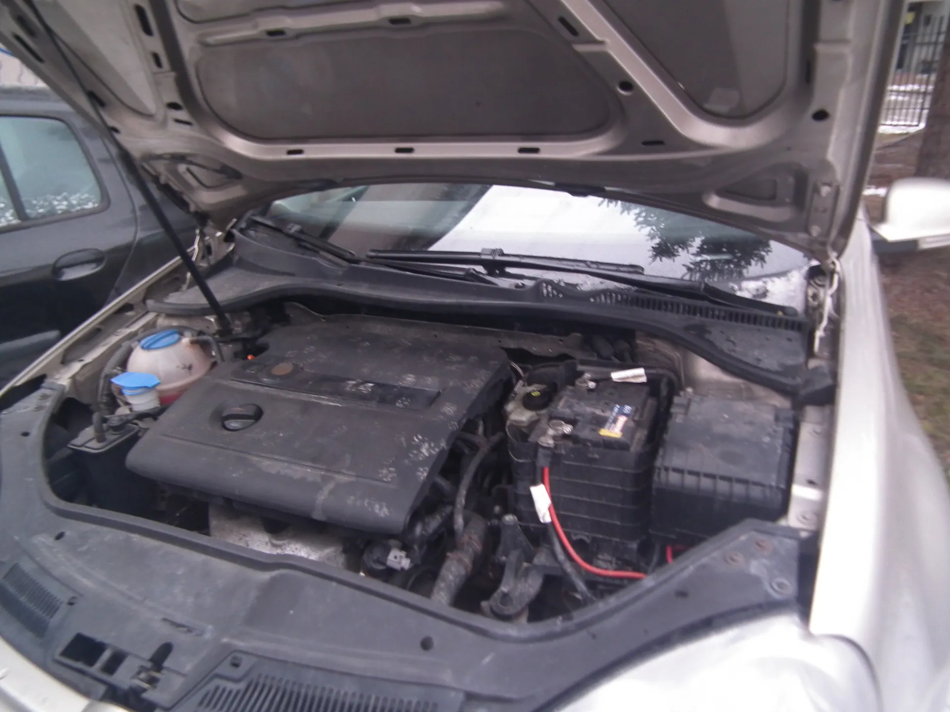 Samochód o srebrnym kolorze z otwartą maską, ukazujący komory silnika. Widać czarną pokrywę silnika, baterię z czerwonymi przewodami, zbiorniki z niebieskimi korkami oraz elementy układu chłodzenia. W tle częściowo widoczny inny samochód i fragment zieleni.