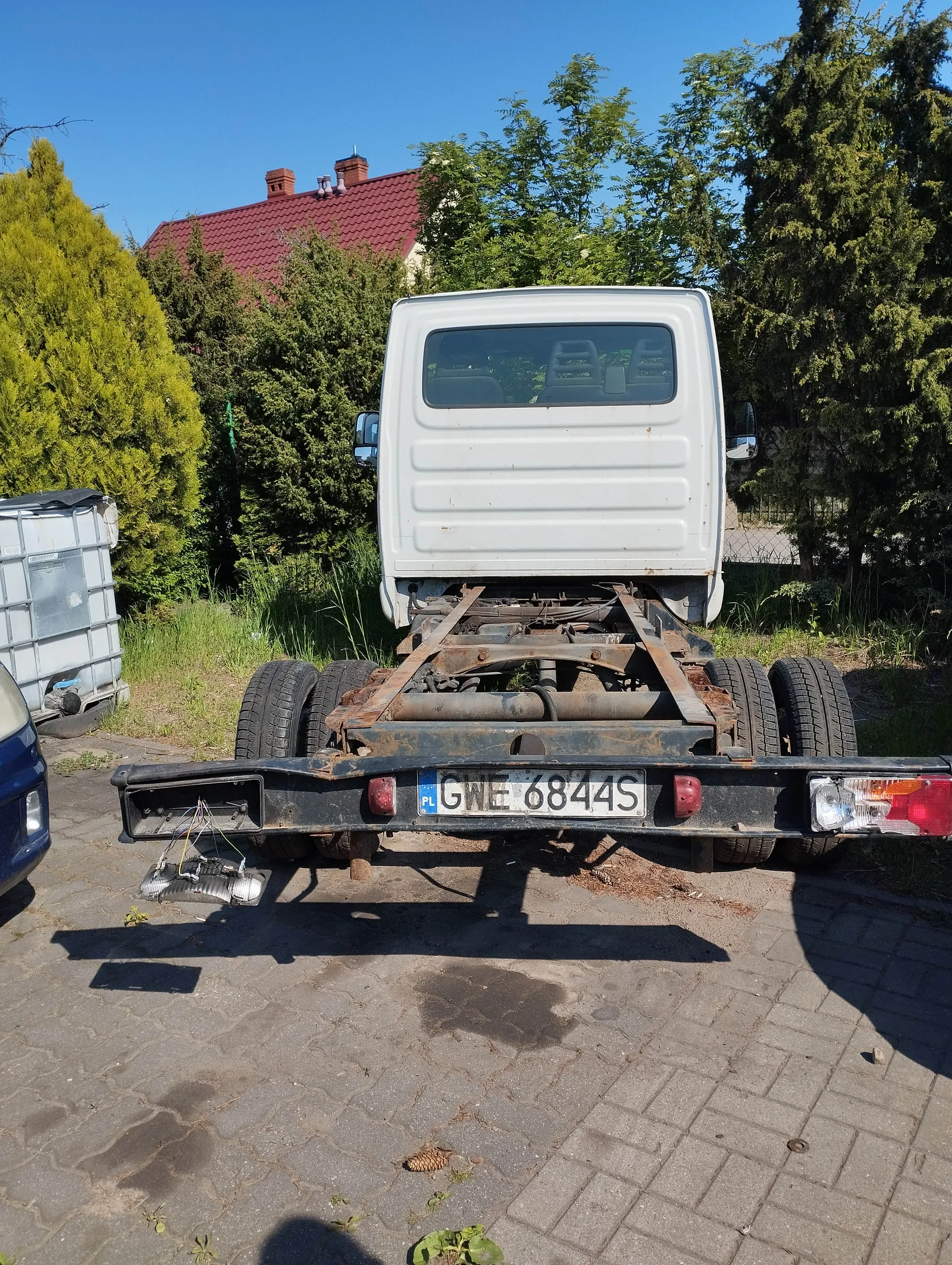 Tył białego samochodu dostawczego bez nadwozia, z widoczną ramą i odsłoniętymi elementami mechanizmu. Na tablicy rejestracyjnej widnieje numer GWE 6844S z oznaczeniem PL po lewej stronie. Samochód stoi na brukowanej powierzchni, w tle zielone krzewy, drzewa oraz dom z czerwonym dachem i ceglanymi kominkami. Po lewej stronie częściowo widoczny niebieski samochód i szary zbiornik. Niebo jest bezchmurne.