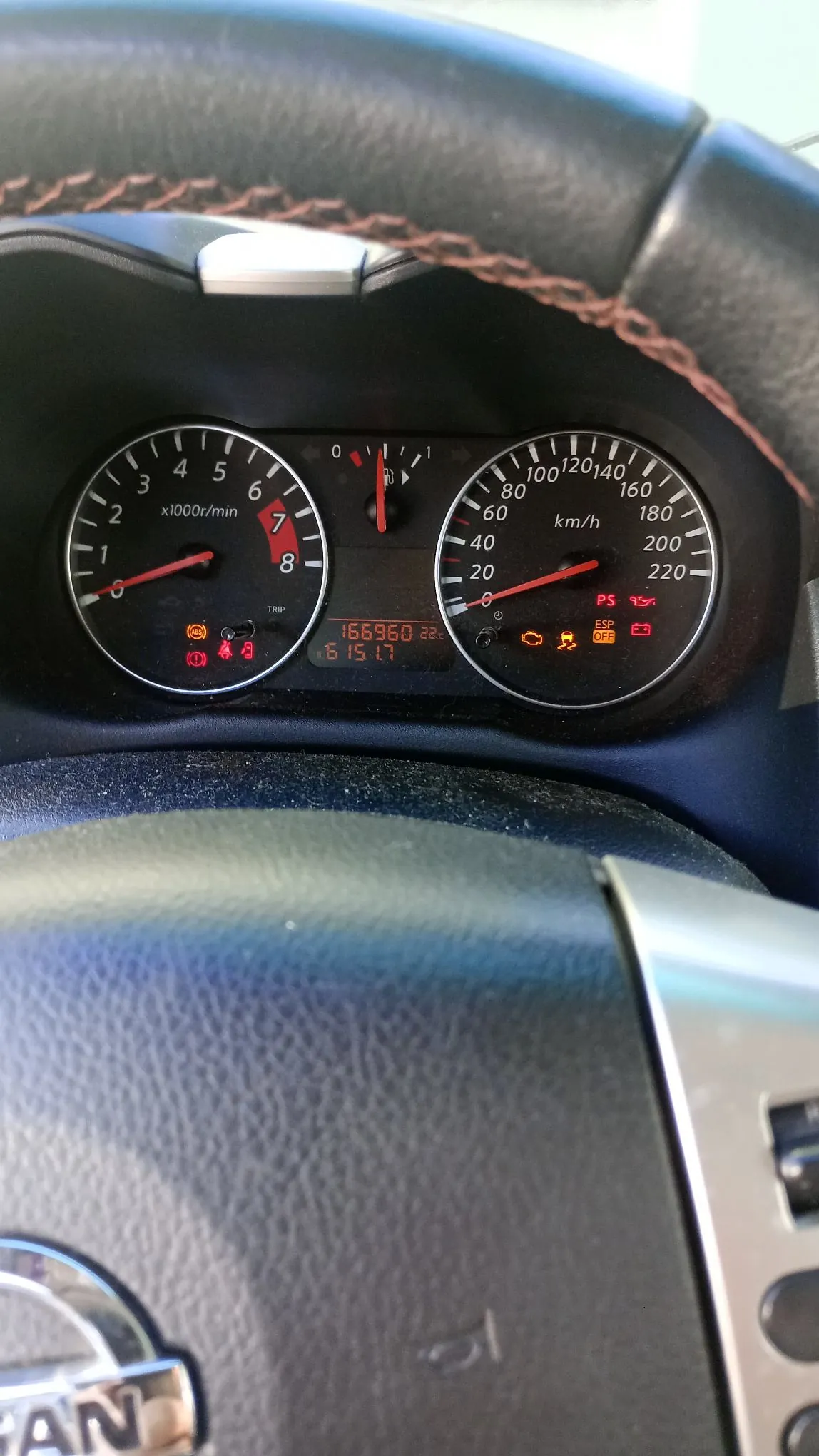 Widok z perspektywy kierowcy na deskę rozdzielczą samochodu marki Nissan. Widoczny jest obrócony fragment kierownicy oraz zestaw wskaźników: po lewej obrotomierz z oznaczeniem x1000r/min, w środku wskaźnik paliwa i temperatury oraz cyfrowy licznik przebiegu wyświetlający wartość 166960, po prawej prędkościomierz z jednostką km/h. Na panelu świecą się kontrolki ostrzegawcze, w tym kontrolka oleju i silnika.