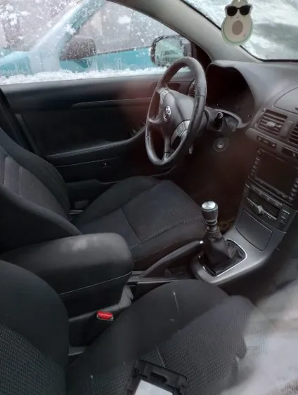 Wnętrze samochodu z widocznym kierownicą po lewej stronie, czarnymi siedzeniami, dźwignią zmiany biegów i deska rozdzielczą. Na lusterku wstecznym wisząca ozdoba w kształcie zielonej twarzy z okularami przeciwsłonecznymi. Za przednią szybą widoczny zasnieżony teren i fragment innego pojazdu. Wnętrze posiada czarne tapicerowane siedzenia, czerwony przycisk na siedzeniu kierowcy oraz radio z klimatyzacją na desce rozdzielczej.