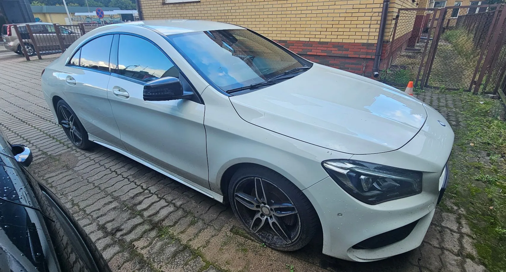 Jasnoszary samochód osobowy typu sedan zaparkowany na kostce brukowej obok budynku z żółtej cegły. Auto jest widoczne z przodu i z boku, ma ciemne felgi aluminiowe. W tle widoczny jest metalowy płot oraz fragment innego, czarnego samochodu po lewej stronie.