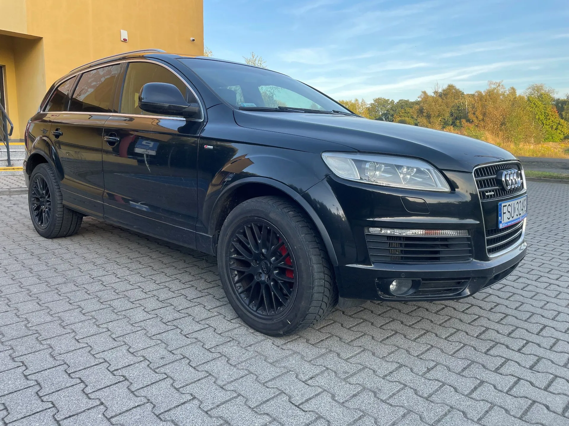 Czarny samochód marki Audi typu SUV zaparkowany na kostce brukowej przed żółtym budynkiem. Pojazd ma czarne felgi, czerwone zaciski hamulcowe i tablicę rejestracyjną o numerze FSU J249. W tle widoczne są drzewa i jasne niebo.