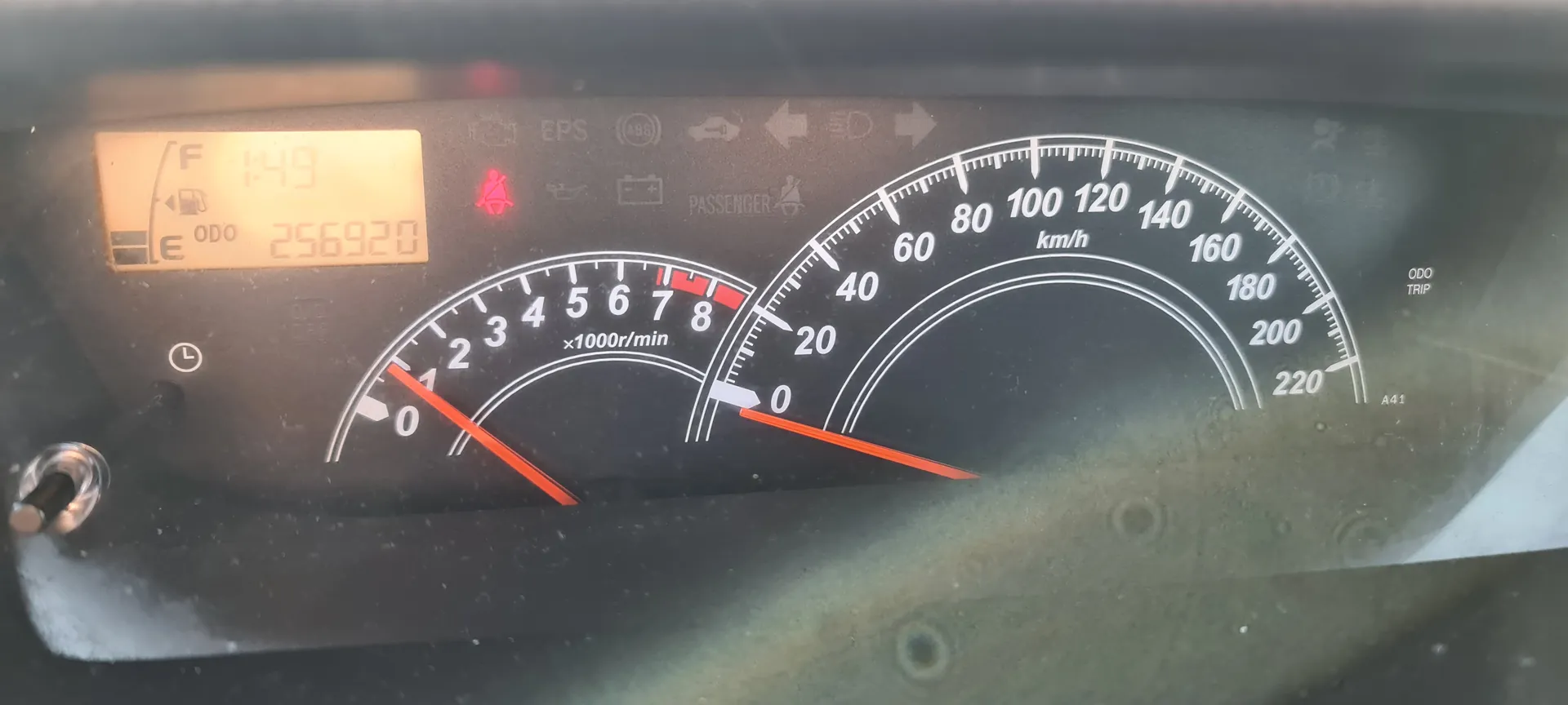 Zbliżenie na tablicę rozdzielczą samochodu. Po lewej stronie wyświetlacz cyfrowy z oznaczeniem paliwa (F), czasem 1:49, licznikiem przebiegu ODO 256920 oraz wskaźnikiem paliwa. W centralnej części znajduje się tachometr (0-8 x1000r/min) z czerwoną igłą wskazującą 0 oraz prędkościomierz (0-220 km/h) z czerwoną igłą wskazującą 0. Widoczne są również ikony ostrzegawcze: czerwona ikona pasów bezpieczeństwa, EPS, ABS, pasażera, baterii i oleju. Na wyświetlaczu widnieją napisy ODO, TRIP oraz A41. Tło tablicy jest ciemne, a znaki i cyfry są białe lub pomarańczowe.