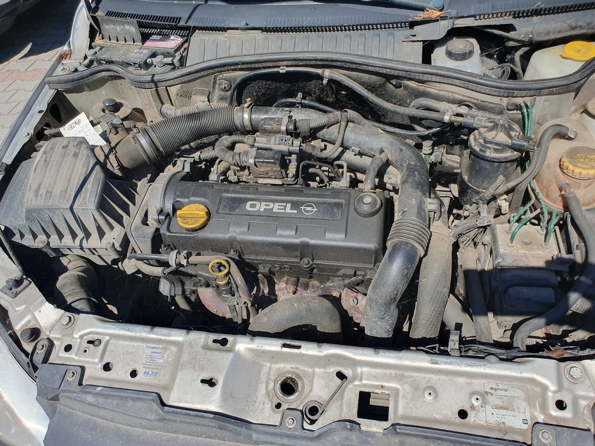Widok z góry na komorę silnika samochodu marki Opel. Widoczna jest czarna pokrywa silnika z napisem 'OPEL' i logo marki, żółty korek wlewu oleju oraz liczne przewody, rury dolotowe i elementy mechaniczne. Silnik jest zakurzony. W dolnej części zdjęcia widoczna jest srebrna rama nadwozia z naklejką techniczną.