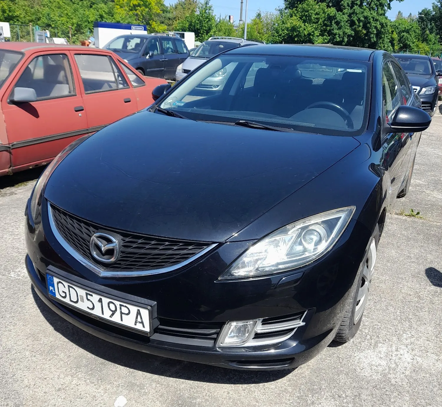 Czarny samochód marki Mazda zaparkowany na betonowym placu. Widoczny jest przód pojazdu z tablicą rejestracyjną o numerze GD 519PA. W tle znajdują się inne samochody, w tym czerwony pojazd po lewej stronie, oraz zielone drzewa.