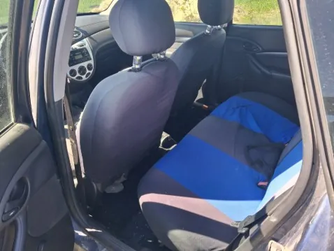 Wnętrze samochodu z otwartą lewą przodem drzwiami. Widoczne są przednie siedzenia: pasażerskie z niebiesko-czarną pokrowcem, kierowcy z ciemnoszarym pokrowcem. W tle deska rozdzielcza z radioodtwarzaczem i wentylacjami, a poza samochodem trawa i fragment drogi. Brak czytelnego tekstu.