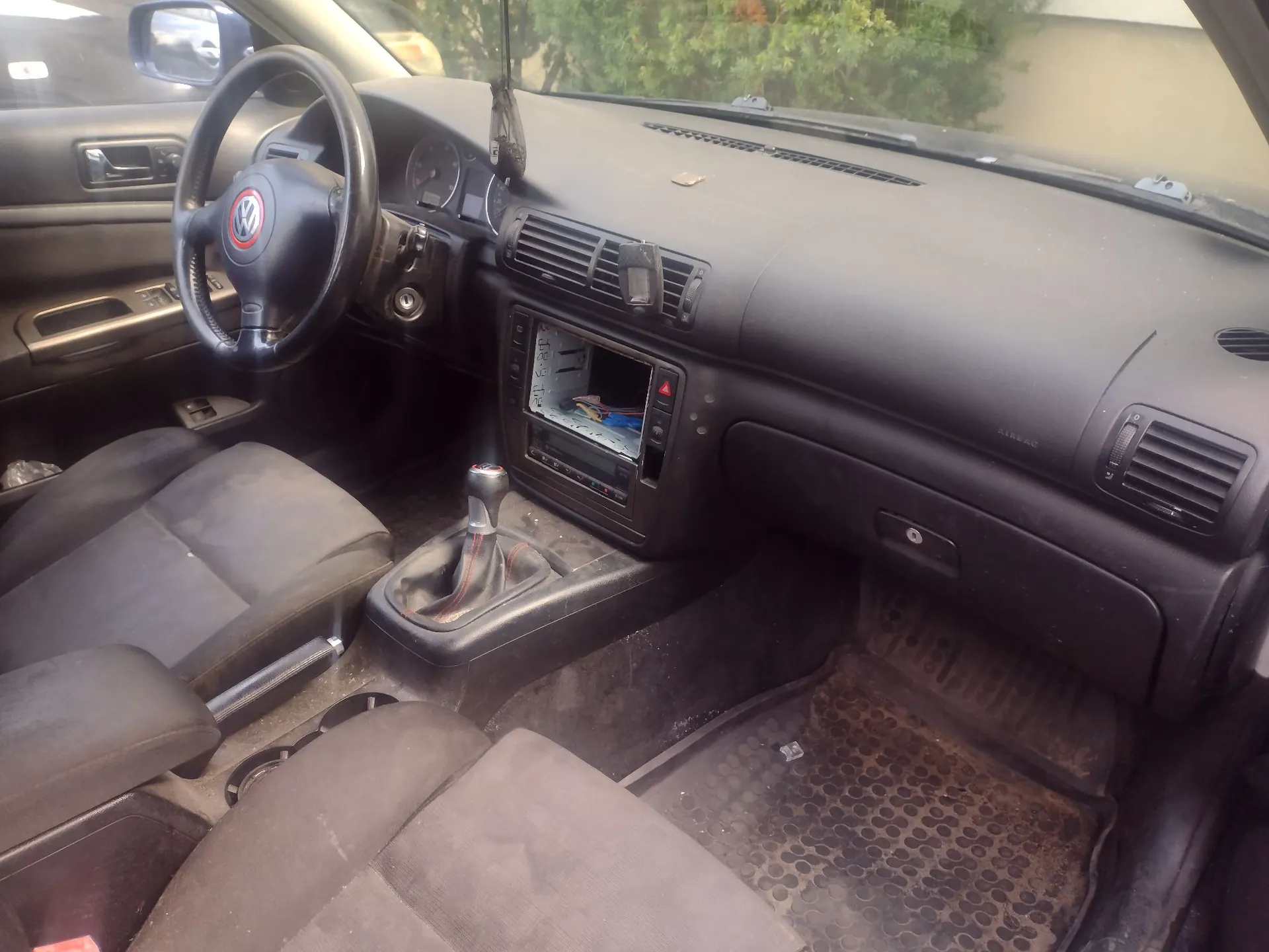 Wnętrze samochodu marki Volkswagen, widok na deskę rozdzielczą, kierownicę z logo VW, konsolę środkową z radiem i drążkiem zmiany biegów oraz przednie fotele z ciemnej tkaniny. Podłoga jest zakurzona i wyłożona gumowymi dywanikami.