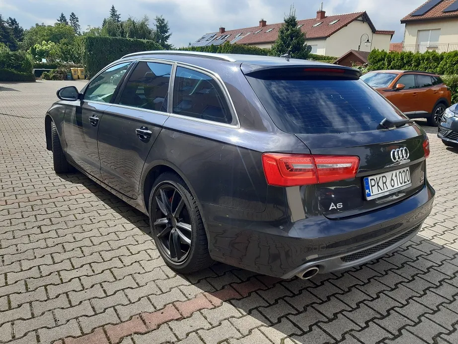 Szary samochód marki Audi A6 w nadwoziu kombi, zaparkowany na kostce brukowej. Widoczny jest tył i bok auta, tablica rejestracyjna o numerze PKR 61020 oraz logo marki. W tle znajdują się domy jednorodzinne, drzewa i inny samochód w kolorze pomarańczowym.