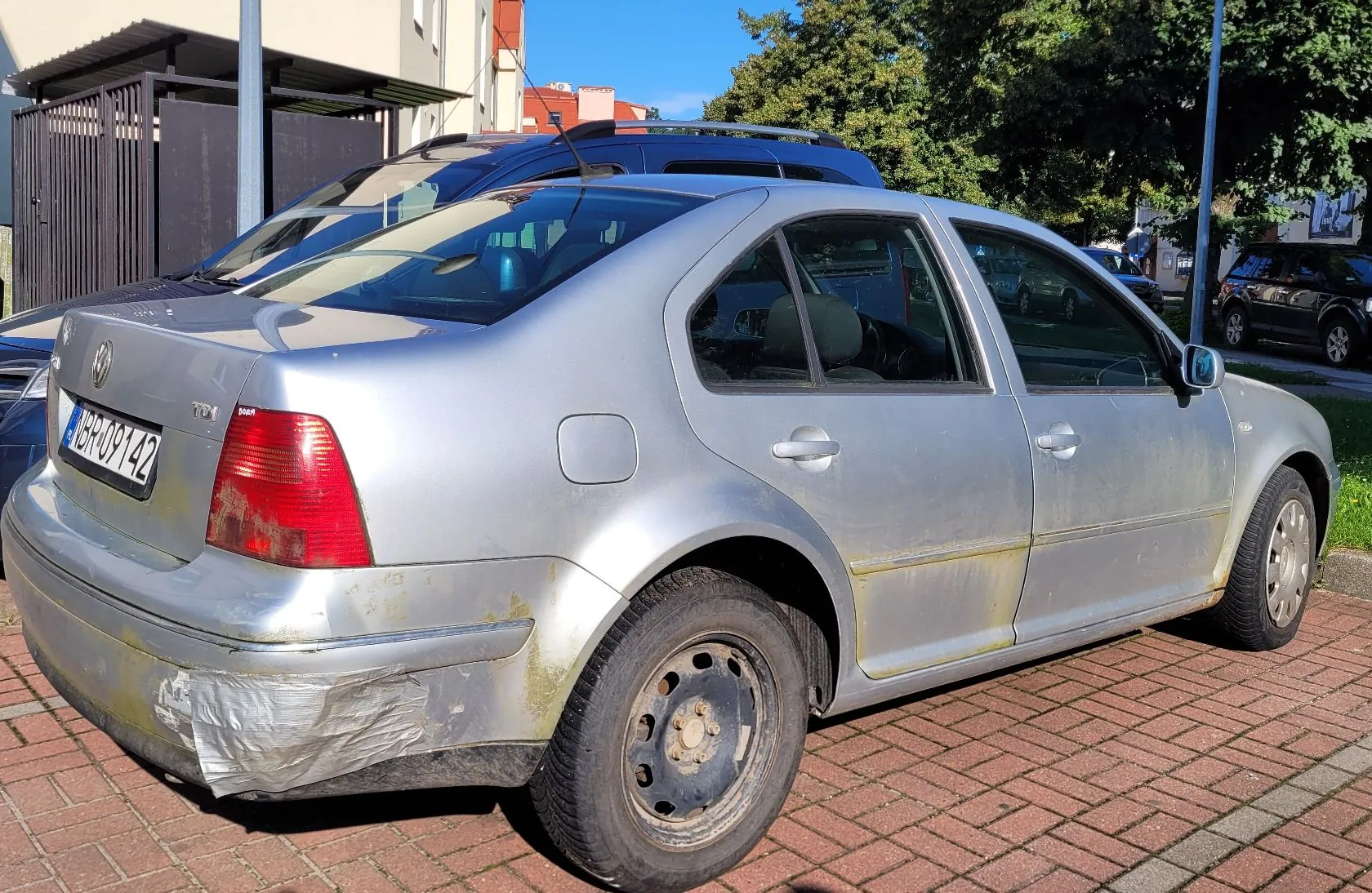Srebrny samochód osobowy marki Volkswagen w nadwoziu sedan, zaparkowany na kostce brukowej. Pojazd jest w złym stanie technicznym: tylny zderzak jest uszkodzony i częściowo zaklejony przezroczystą taśmą, a na dolnych krawędziach nadwozia widoczne są ślady rdzy. Samochód posiada tablicę rejestracyjną o numerze ABR 09142. W tle widoczne są inne zaparkowane samochody, budynek oraz drzewa.