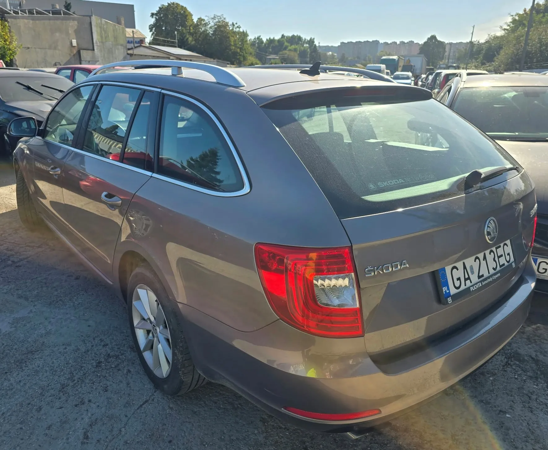Szary samochód marki Skoda w nadwoziu kombi, zaparkowany na placu obok innych pojazdów. Widoczny jest tył i lewy bok auta. Samochód posiada srebrne relingi dachowe i tablicę rejestracyjną o numerze GA 213EG. Na klapie bagażnika znajduje się napis ŠKODA oraz logo marki.