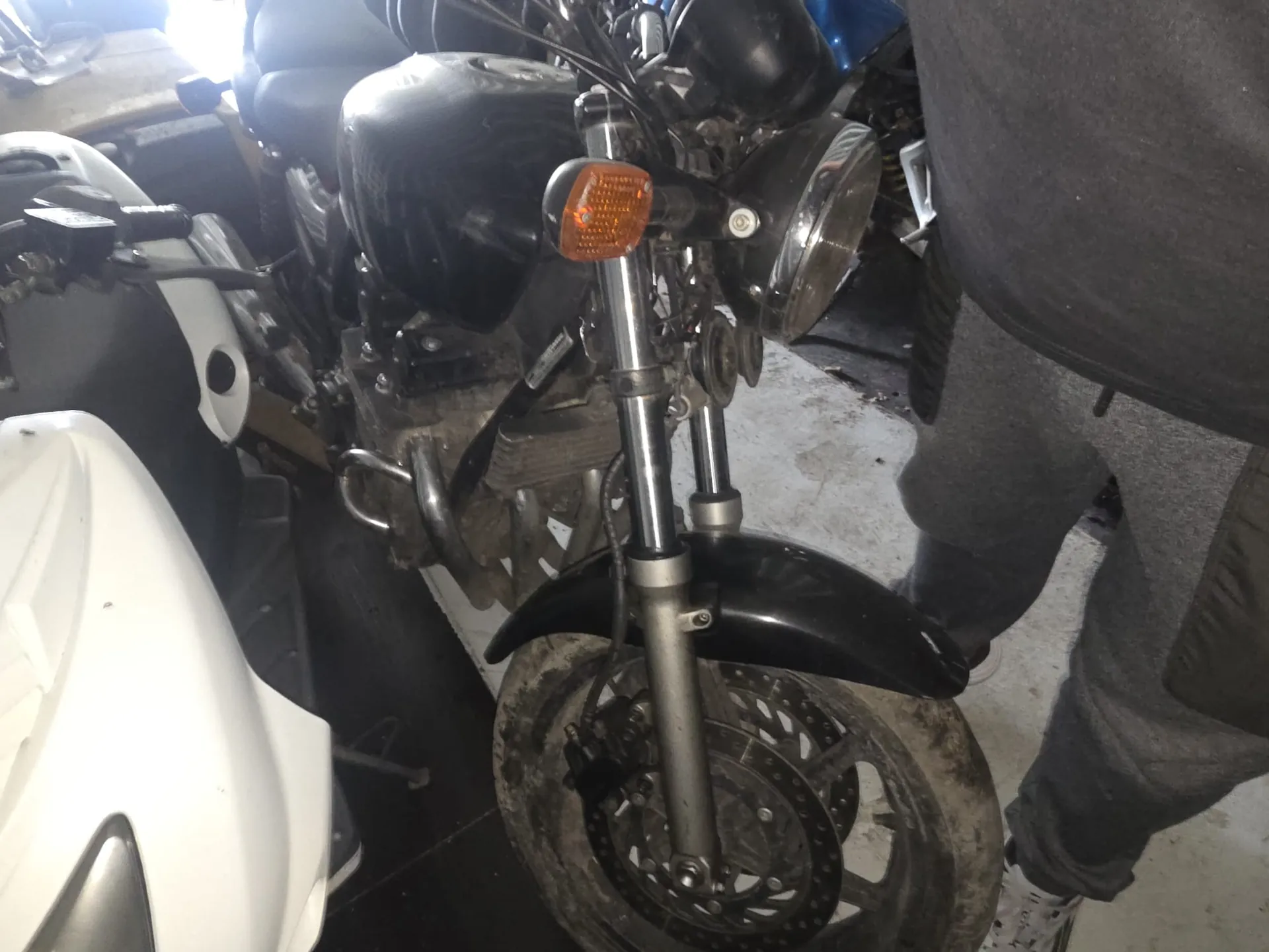 Zbliżenie na przednią część czarnego motocykla z widocznym reflektorem, pomarańczowym kierunkowskazem, czarnym zbiornikiem paliwa i brudnym kołem przednim. Po prawej stronie widoczna jest osoba w szarych spodniach i białych butach stojąca obok motocykla. W tle częściowo widoczny biały pojazd oraz betonowa podłoga garażu.