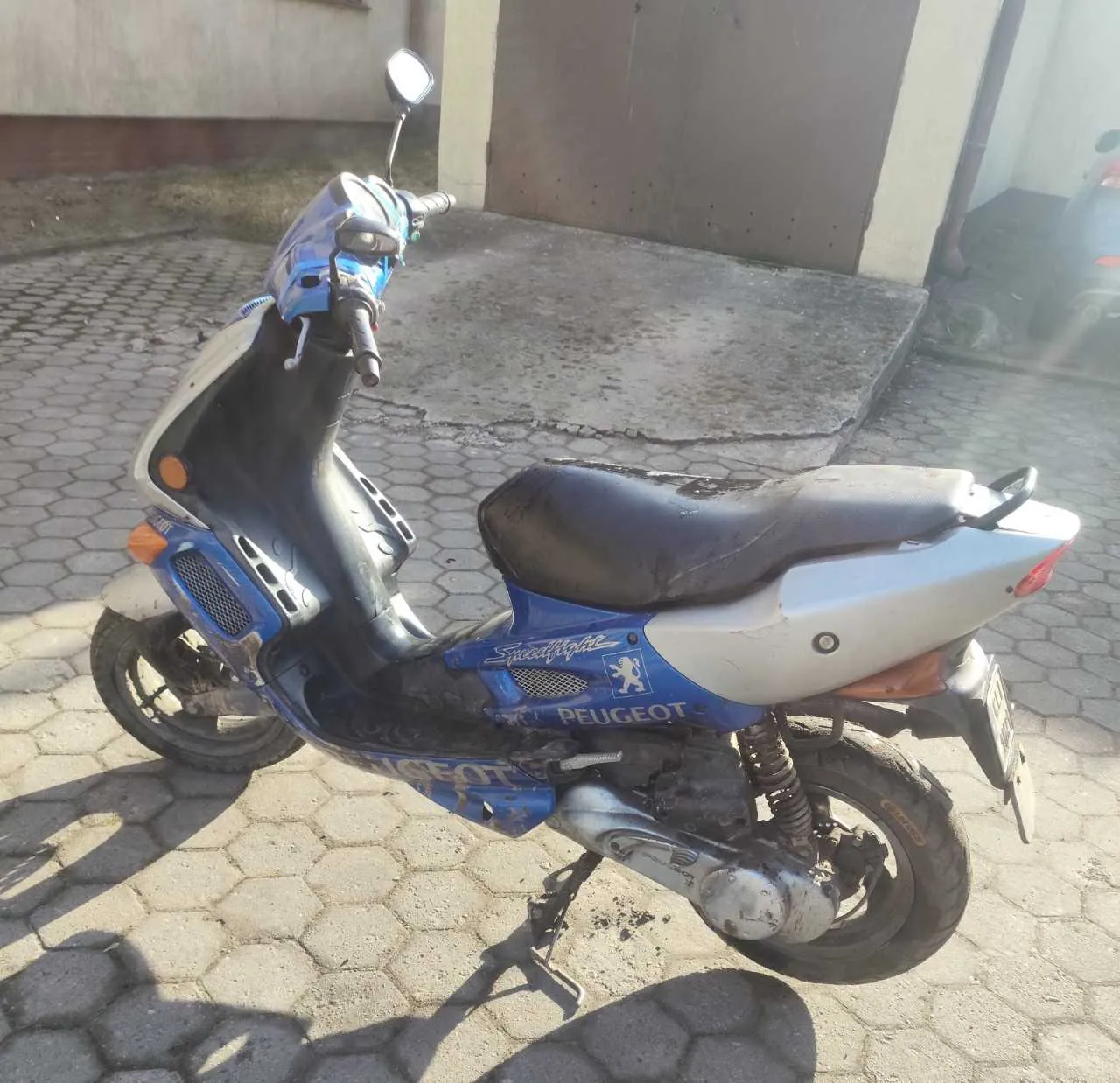 Niebiesko-biały skuter marki Peugeot model Speedfight zaparkowany na chodniku z sześciokątnymi płytami. Skuter ma czarne siedzenie, widoczne logo Peugeot oraz napis Speedfight na boku. W tle znajduje się betonowa ściana i brązowe drzwi garażowe. Na skuterze widoczne są ślady zużycia i brudu.