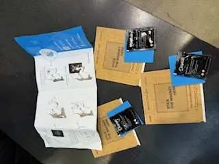 Na ciemnej powierzchni leżą różne przedmioty: dwa niebieskie koperty lub folderów, kilka brązowych kopert papierowych, biały składany dokument z ilustracjami oraz trzy małe czarne opakowania z niebieską taśmą. Wszystkie elementy są rozmieszczone w sposób nieuporządkowany.