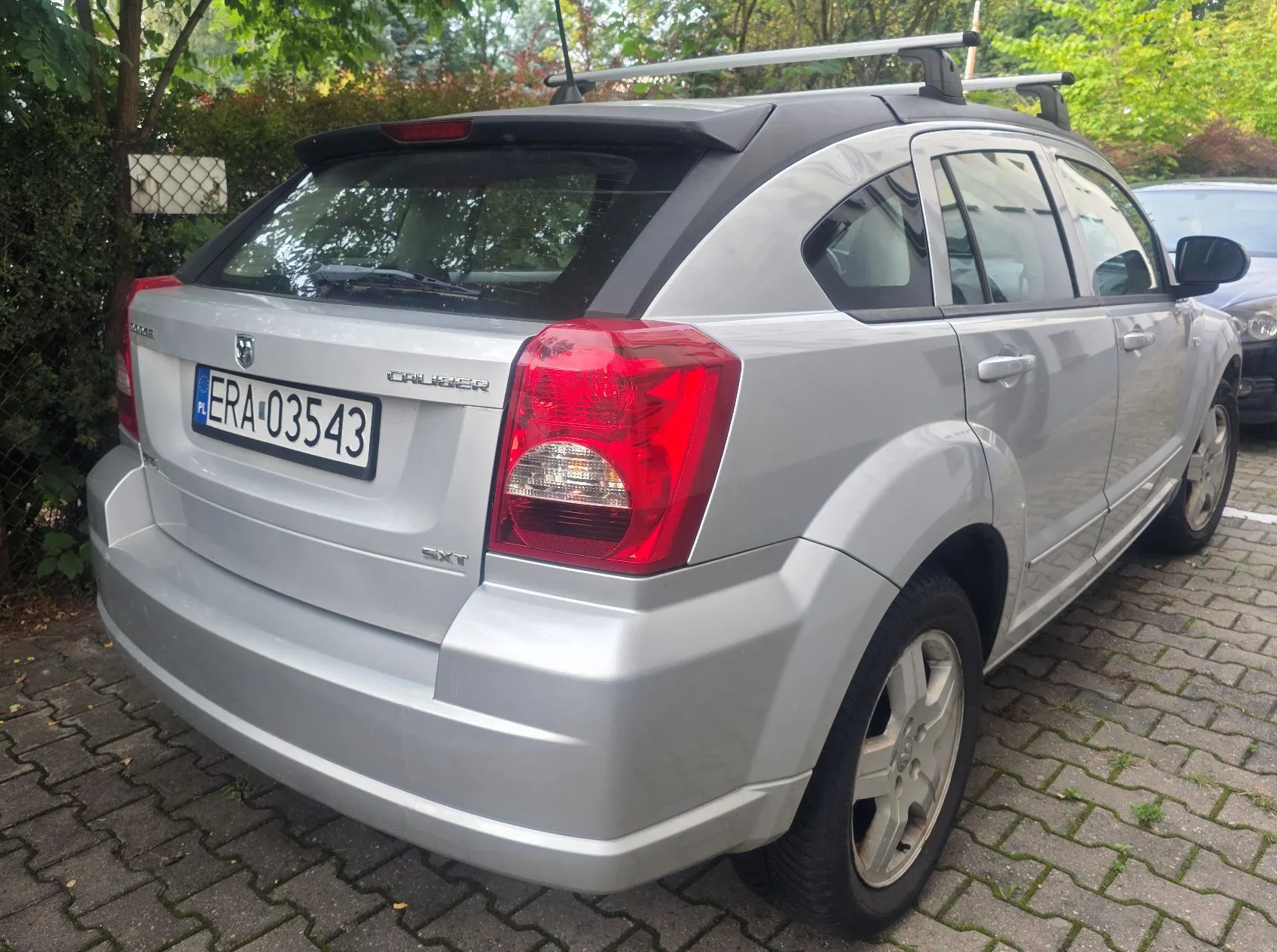 Srebrny samochód marki Dodge Caliber w wersji SXT, zaparkowany na kostce brukowej. Widoczny jest tył pojazdu z tablicą rejestracyjną ERA 03543, bagażnikiem dachowym oraz logo Dodge i napisem Caliber na klapie bagażnika.