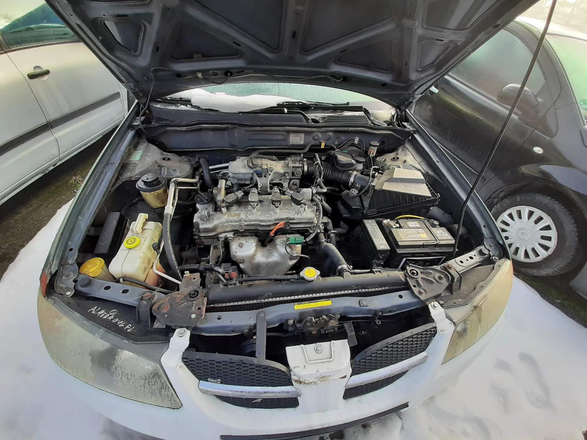 Samochód z otwartą maską, ukazujący komorę silnika. Widoczne są elementy takie jak akumulator, zbiorniki płynów z żółtymi korkami, silnik oraz różne przewody. Na przedniej części komory silnika znajduje się napis 'AL4 1.4 16V'. Samochód jest zaparkowany na śniegu, obok innych pojazdów – srebrnego po lewej i czarnego po prawej stronie. Tło obejmuje fragmenty innych samochodów i ziemię z resztkami śniegu.