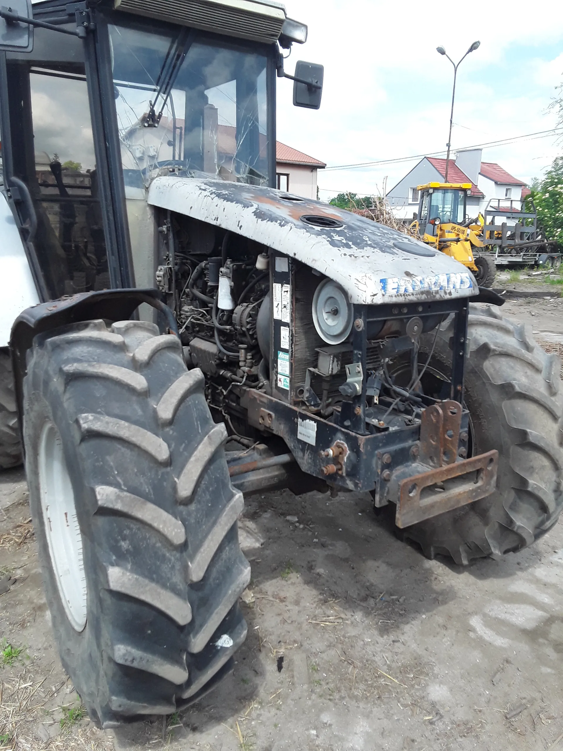 Traktor z dużymi oponami, częścią silnika i kabiną z szybą. Na białej części traktora widoczny napis 'FARMER'. W tle żółty pojazd budowlany, domy z czerwonymi dachami, latarnia uliczna oraz niebo z chmurami. Na ziemi sucha trawa i ślad po oponach.