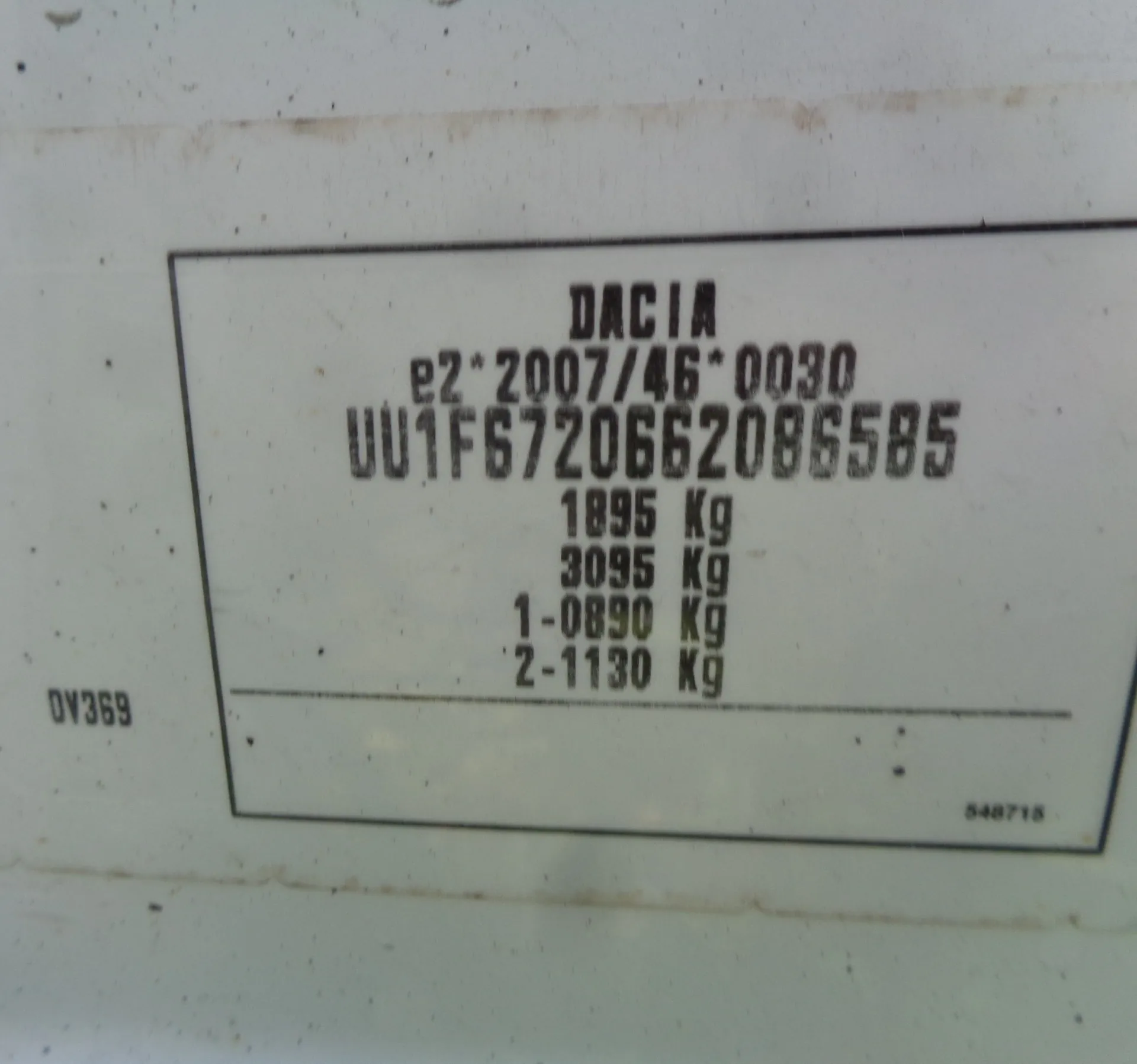 Zdjęcie tabliczki znamionowej pojazdu marki DACIA. Na białym tle w czarnej ramce znajduje się czarny tekst: 'DACIA', poniżej 'e2*2007/46*0030', następnie numer seryjny 'UU1F6720662086585'. Poniżej widnieją wartości masy: '1895 Kg', '3095 Kg', '1-0890 Kg' oraz '2-1130 Kg'. Po lewej stronie ramki widnieje napis 'DV369', a w prawym dolnym rogu wewnątrz ramki liczba '548715'.