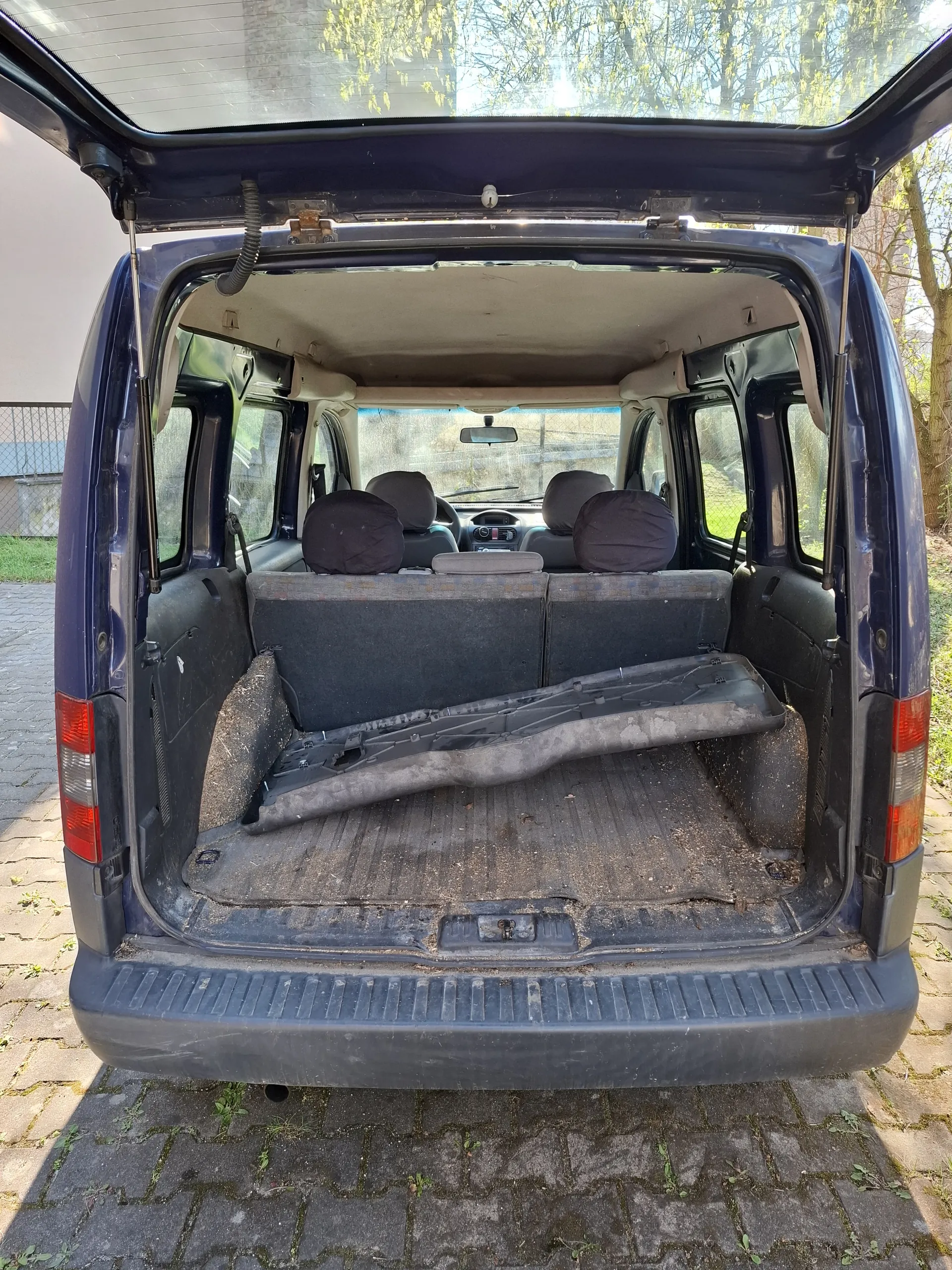 Ciemnoniebieski samochód dostawczy z otwartą tylną klapą. Wnętrze z widocznymi siedzeniami, część z nich złożona. Podłoga bagażnika zabrudzona. Samochód zaparkowany na brukowanej powierzchni, w tle drzewa i ogrodzenie.