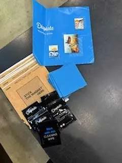 Niebieska teczka z logo 'Dunkin'' i małym zdjęciem, stos niebieskich kartek, brązowe pudełko z napisem 'PRIORITY MAIL' oraz kilka czarnych opakowań z widocznymi napisami 'DARK' i 'MEDIUM' ułożonych na szarym tle.