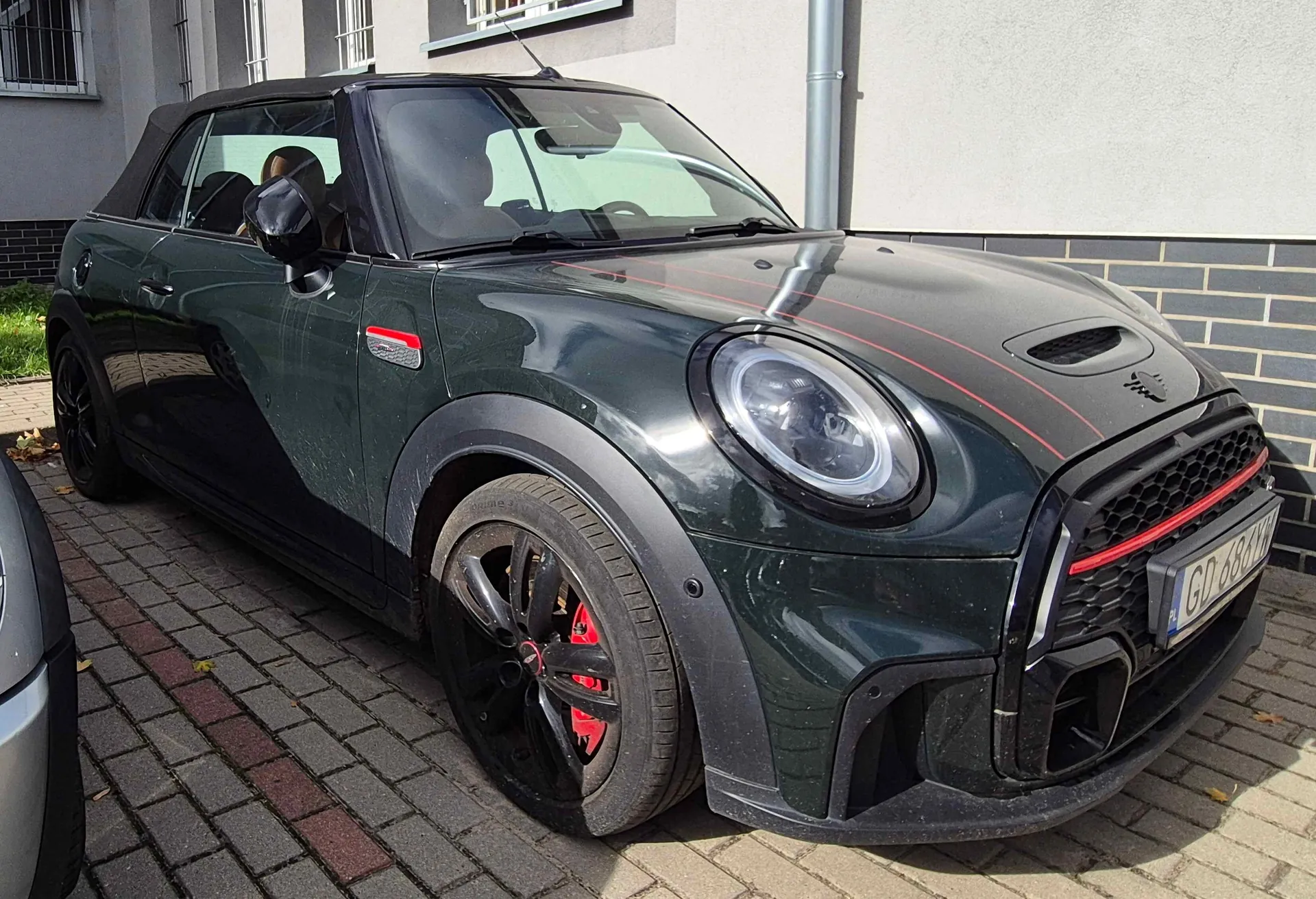 Ciemnozielony samochód Mini Cooper Convertible zaparkowany na kostce brukowej przed jasnym budynkiem. Auto ma czarny miękki dach, czarne felgi z czerwonymi zaciskami hamulcowymi oraz czerwone detale na masce i grillu. Widoczna jest polska tablica rejestracyjna GD 680Y.