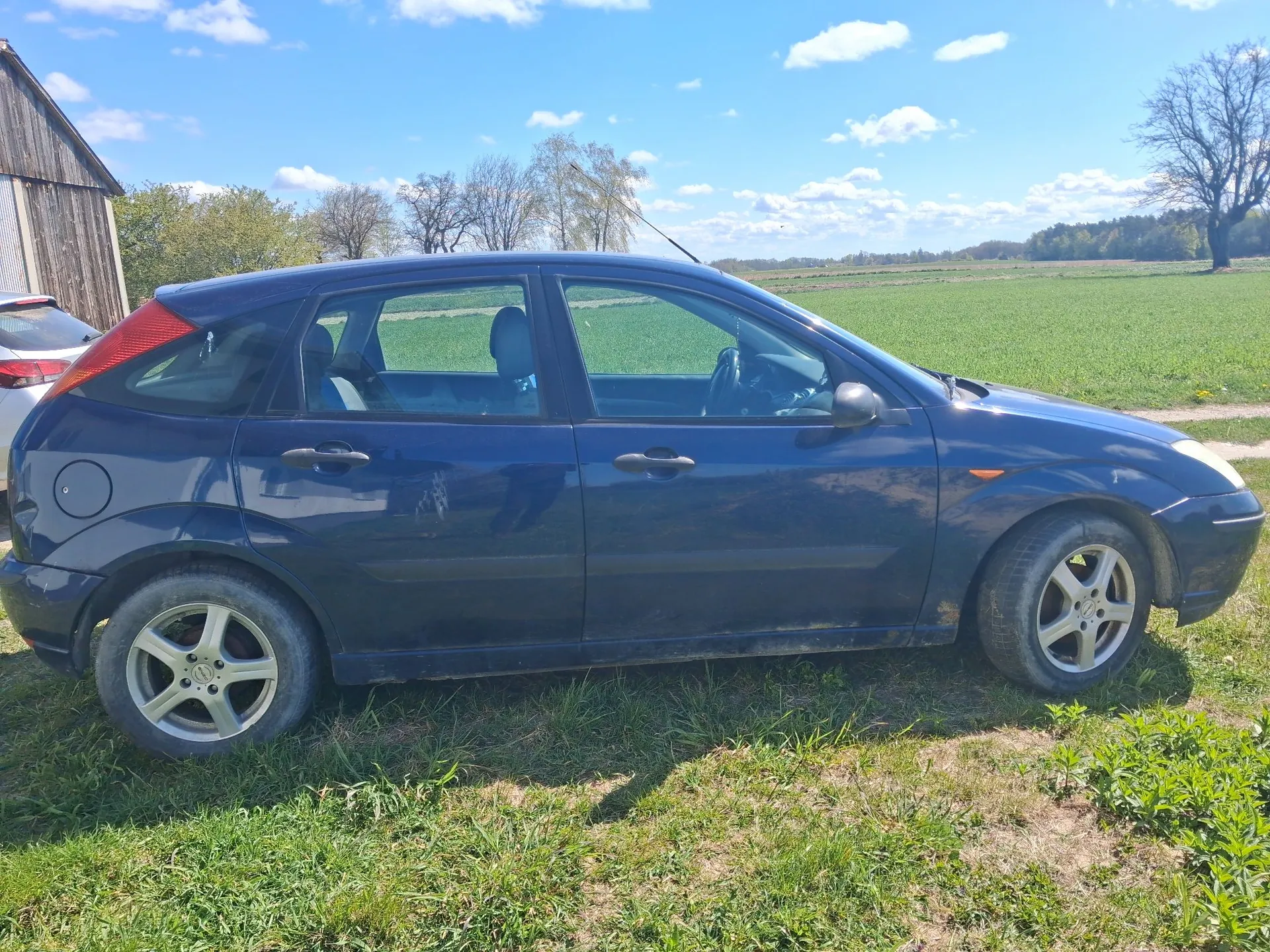 Granatowy samochód osobowy typu hatchback zaparkowany na trawiastym terenie. Pojazd jest pokazany z profilu, z boku. W tle widać zielone pole, kilka drzew oraz fragment drewnianego budynku po lewej stronie pod jasnoniebieskim niebem z białymi chmurami.