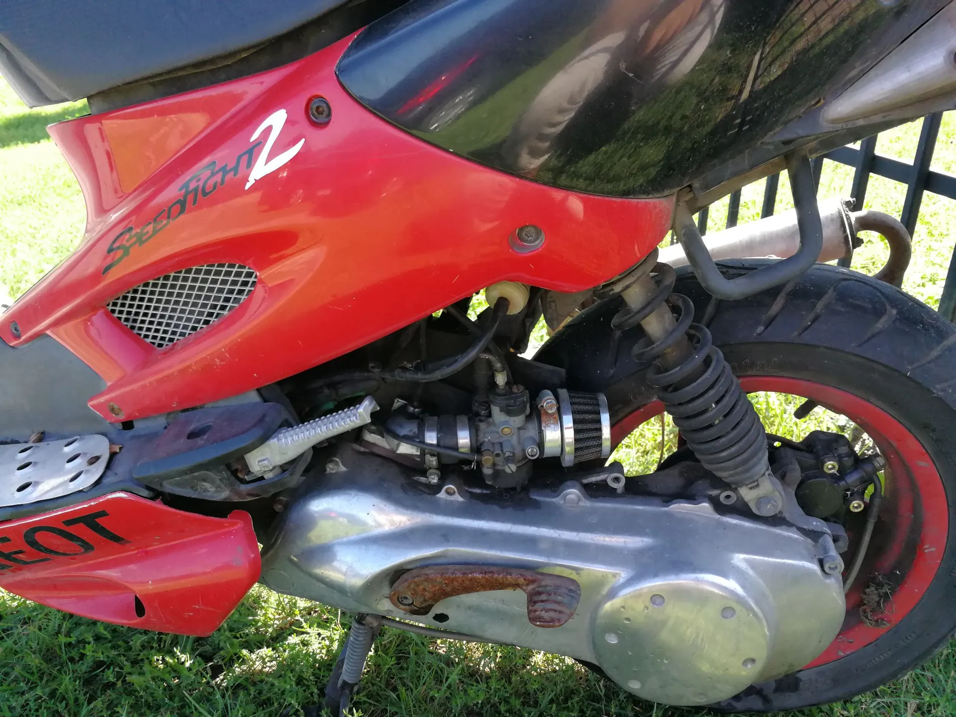 Zbliżenie na prawą stronę czerwonego skutera. Na czerwonej owiewce widnieje biały napis „SPEEDFIGHT 2”. Widoczne są elementy mechaniczne: srebrna obudowa przekładni, amortyzator, czarna opona na czerwonej feldze oraz fragment rury wydechowej. Skuter stoi na trawie.