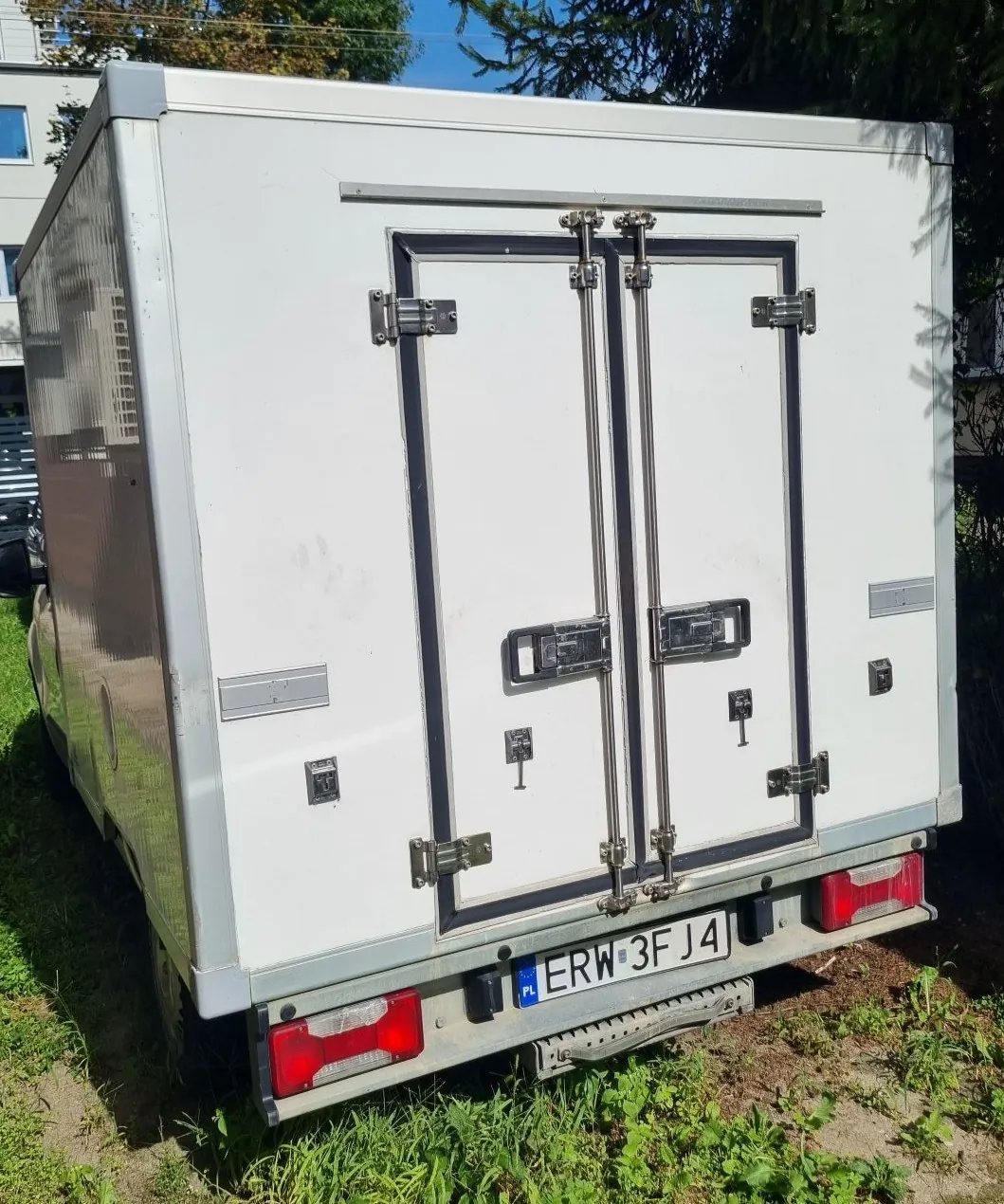 Tylny widok białej furgonetki z zabudową kontenerową. Pojazd posiada dwuskrzydłowe drzwi zamykane na rygle i zawiasy. Na zderzaku znajduje się polska tablica rejestracyjna o numerze ERW 3FJ4 oraz dwa czerwone światła tylne. Pojazd stoi na trawiastym podłożu.