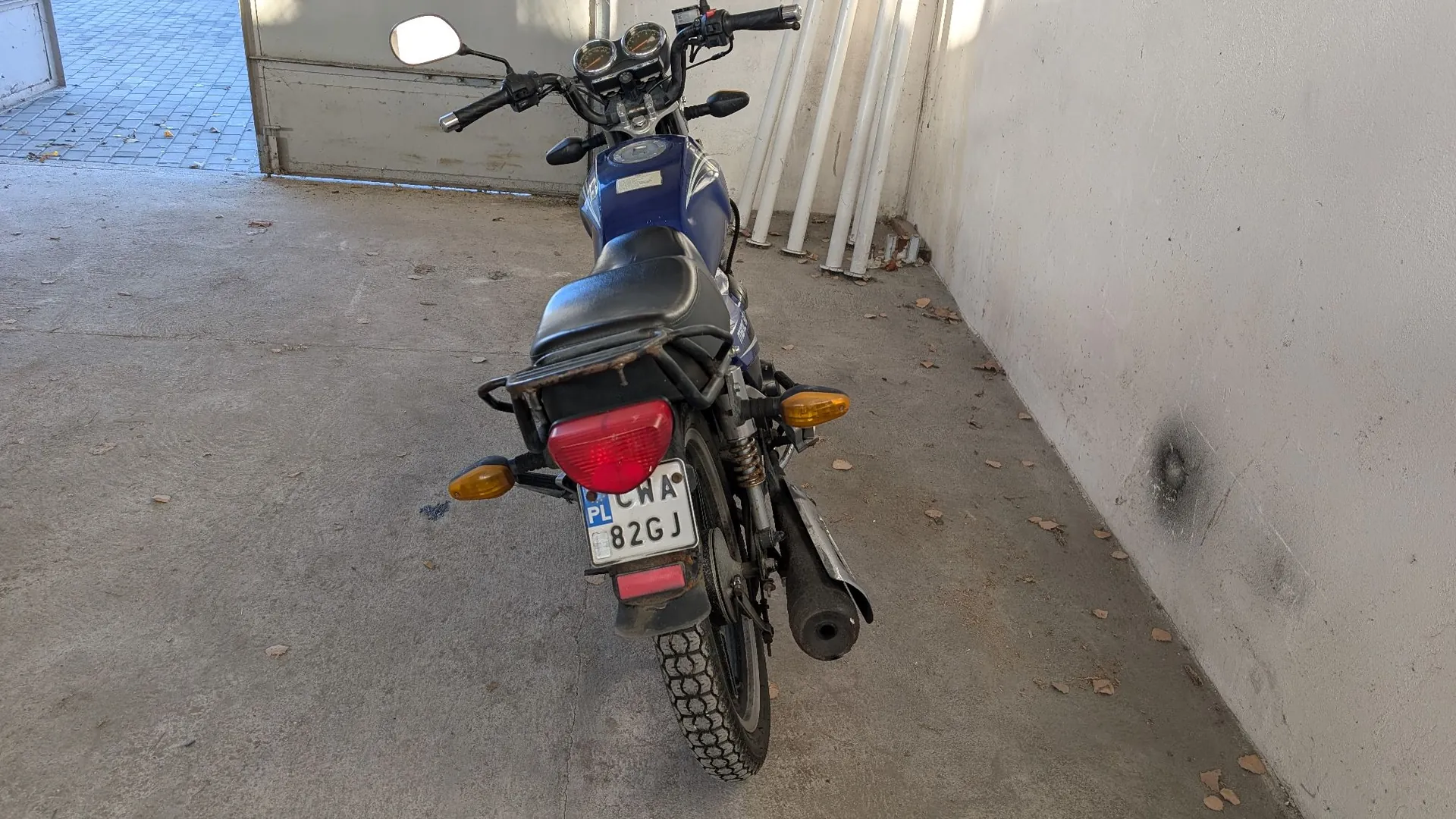 Niebieski motocykl zaparkowany tyłem do widza na betonowym podłożu obok białej ściany. Motocykl posiada czarne siedzenie, tylne światło stopu oraz polską tablicę rejestracyjną o numerze WA 82GJ. Po prawej stronie widoczna jest biała ściana z kilkoma rurami przytwierdzonymi do niej.