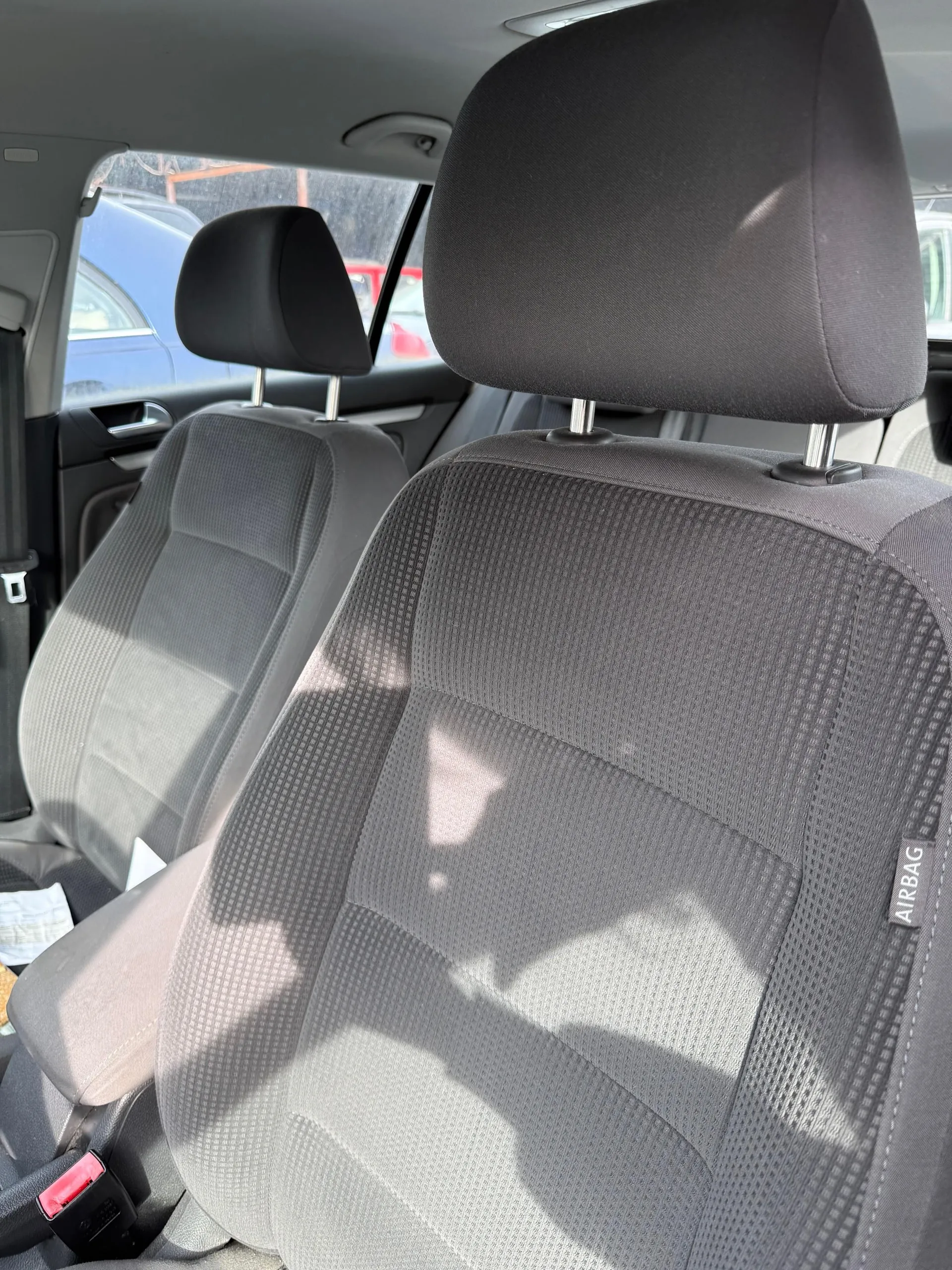 Wnętrze samochodu z widocznymi przodowymi siedzeniami w kolorze szarym o teksturze siateczkowej. Na prawym siedzeniu widoczny etykietka z napisem AIRBAG. Widoczne poduszki głowowe, pasy bezpieczeństwa z czerwonym przyciskiem oraz fragmenty zewnętrznych pojazdów (niebieski i czerwony samochód) przez okno. Oświetlenie słoneczne rzuca cienie na siedzenia.