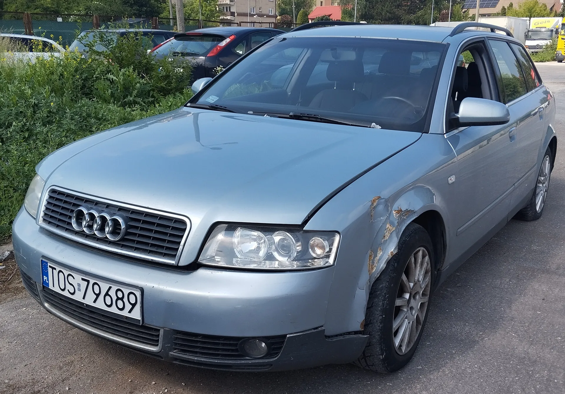 Srebrny samochód Audi typu kombi zaparkowany na asfaltowej powierzchni. Widoczne uszkodzenie z rdzą na prawym przednim błotniku. Na przodzie tablica rejestracyjna z polskim kodem PL i numerem TOS 79689. W tle inne pojazdy, zielone krzewy, ogrodzenie i budynki. Logo Audi na atrapie chłodnicy. Tekst na tablicy rejestracyjnej jest czytelny i poziomo ustawiony.