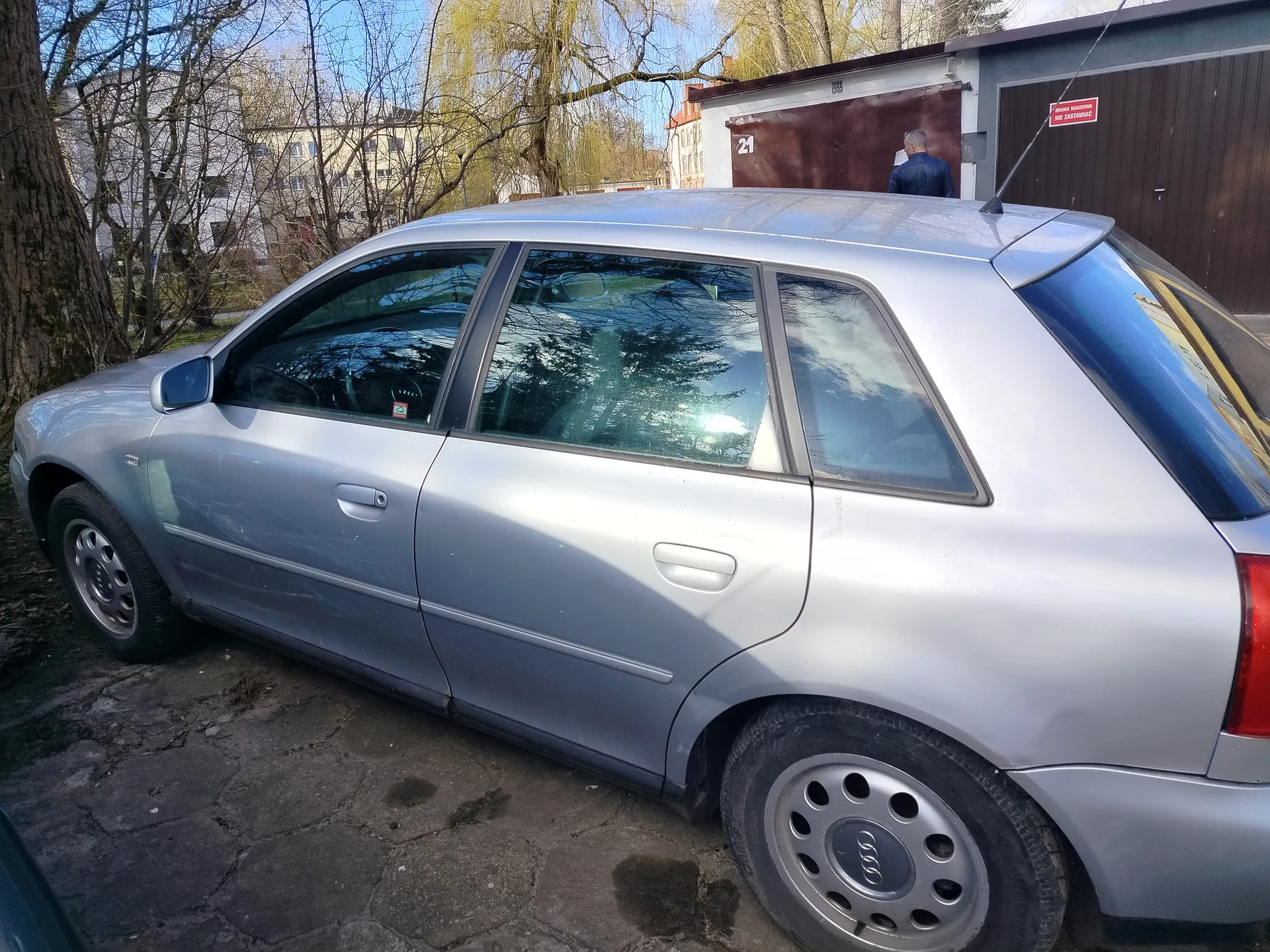 Srebrny samochód Audi typu hatchback zaparkowany na brukowanej powierzchni. W tle drzewa bez liści, budynki mieszkalne oraz garaż z brązowymi drzwiami. Na ścianie garażu widoczny biały numer "21" oraz czerwony znak z białym napisem "ZABRONIONY WJAZD". Przy garażu stoi mężczyzna w ciemnej kurtce. Niebo jest błękitne, a na ziemi widoczne są plamy wilgoci.