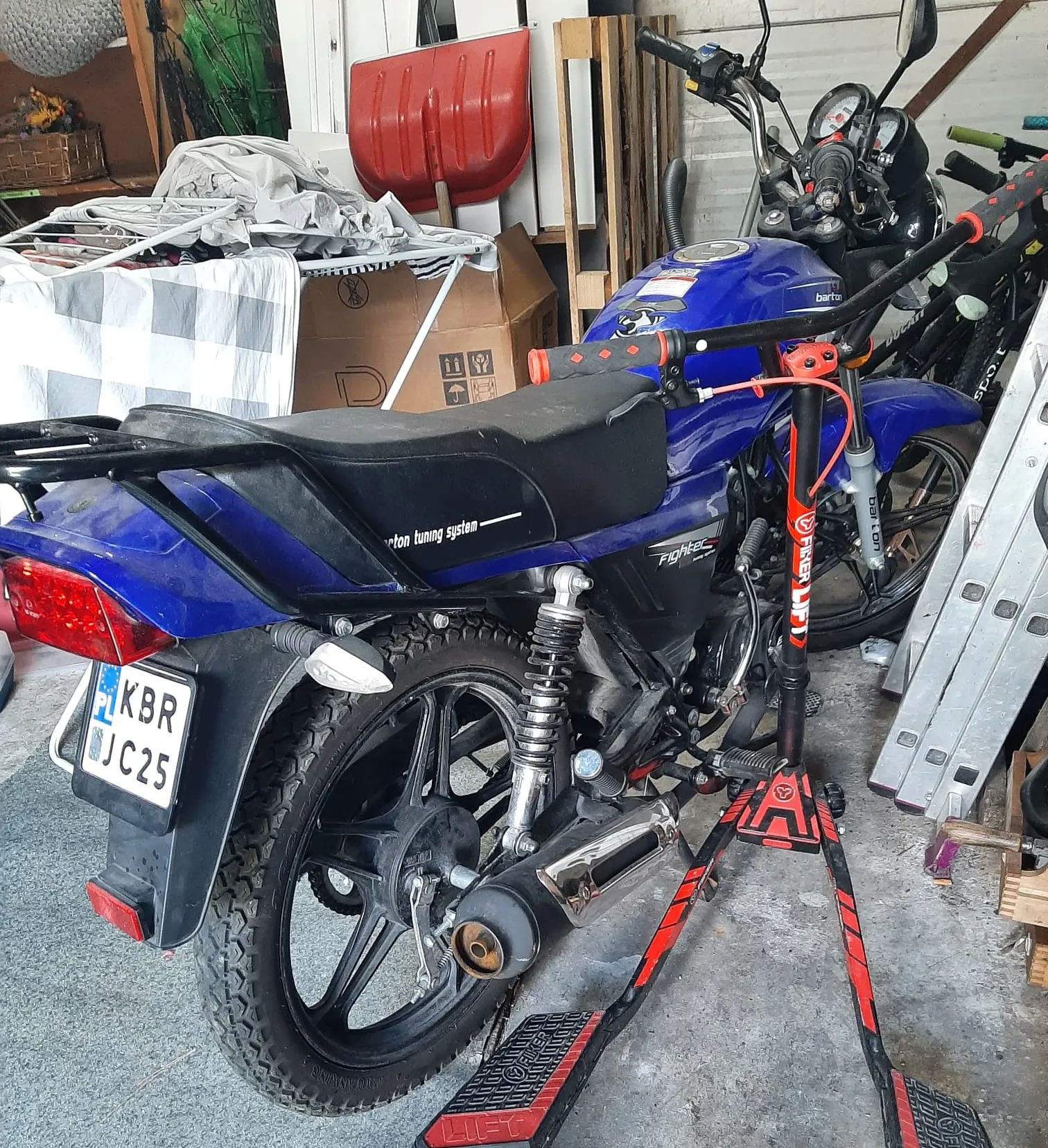 Niebieski motocykl marki Bartor model Fighter zaparkowany w garażu na czerwono-czarnym podstawie. Widoczna tablica rejestracyjna PL KBR JC25, czarne siedzenie z napisem 'borton tuning system', czerwone światło tylnego reflektora oraz wydech. W tle: czerwona łopata, kartony, biało-szara kratowana tkanina, drewniane deski i części rowerów. Na motocyklu widoczne elementy jak wskaźniki, czarne koło z grubą oponą oraz czerwone przewody.