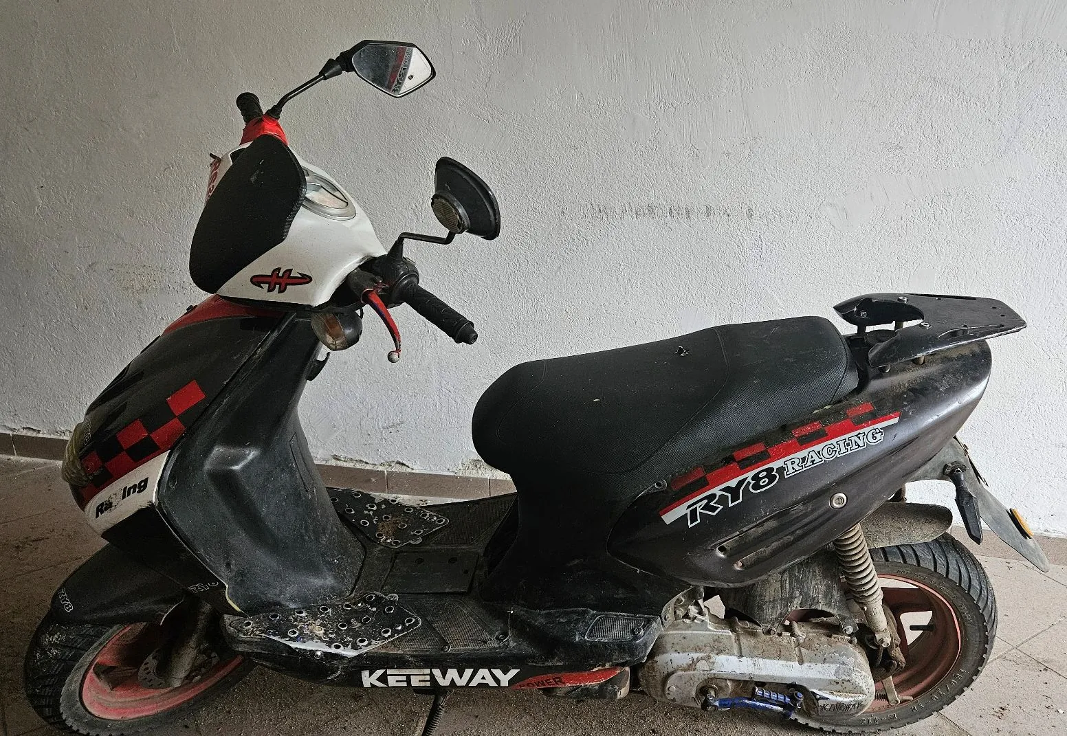 Czarny skuter marki Keeway z białymi i czerwonymi akcentami. Na boku siedzenia znajduje się napis 'KY8 RACING', a na dolnej części ramy napis 'KEEWAY'. Pojazd ma dwa lusterka, czarne siedzenie i czerwone felgi. Stoi na jasnej, betonowej powierzchni na tle białej ściany.