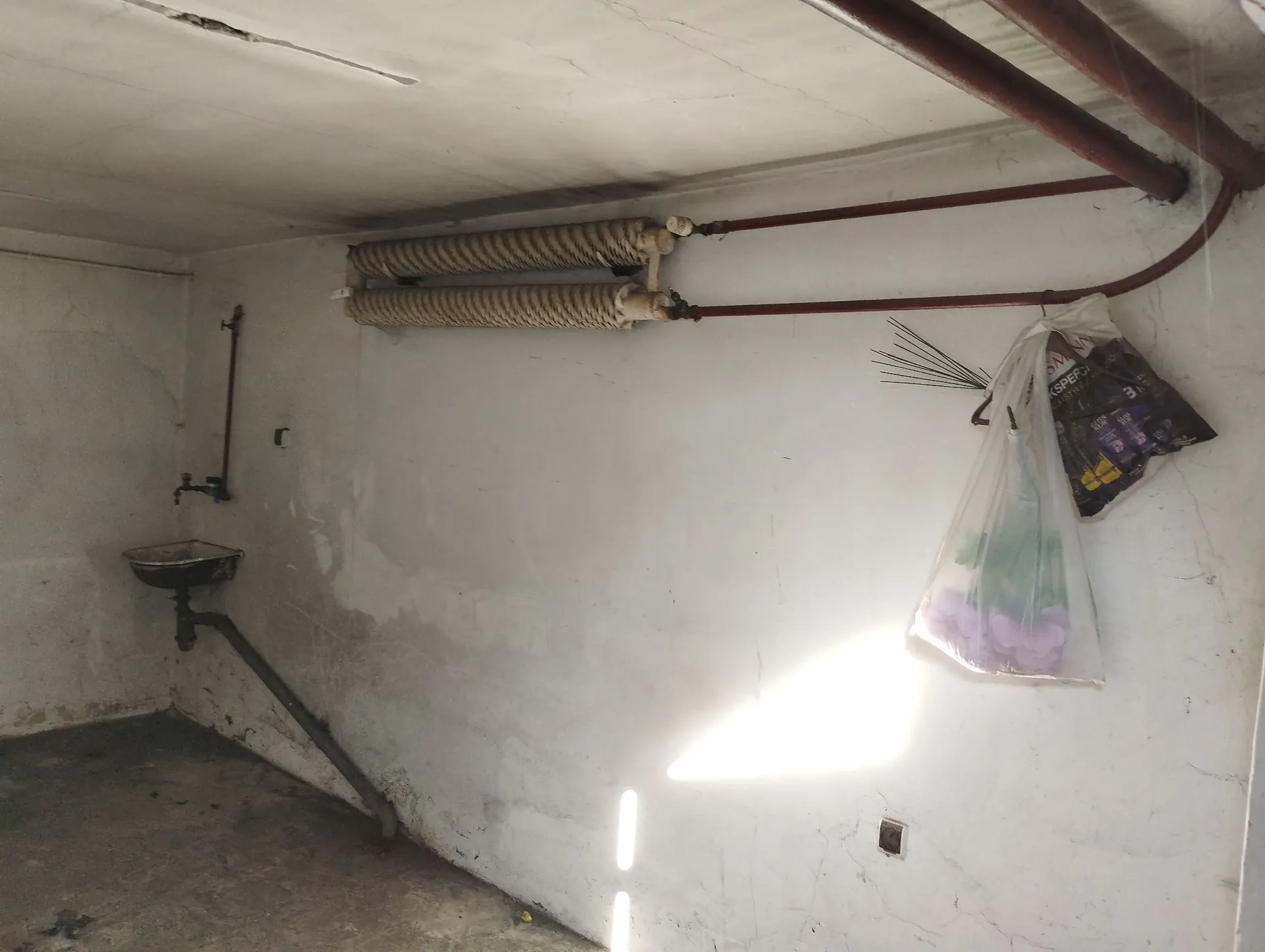 Wnętrze pomieszczenia z białymi, zabrudzonymi ścianami i betonową podłogą. Po lewej stronie znajduje się mały, metalowy zlew z rurą odpływową biegnącą wzdłuż ściany. Na ścianie zamontowane są dwa poziome, żebrowane grzejniki połączone czerwonymi rurami, które biegną wzdłuż sufitu. Po prawej stronie wisi przezroczysta plastikowa torba z fioletowymi kwiatami i ciemnym opakowaniem.
