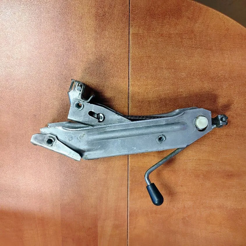 Metalowy mechanizm, prawdopodobnie część samochodowa, leżący na brązowym, drewnianym blacie. Element składa się z szarego, odlewanego korpusu z widoczną sprężyną oraz metalowej dźwigni zakończonej czarną, plastikową rączką.
