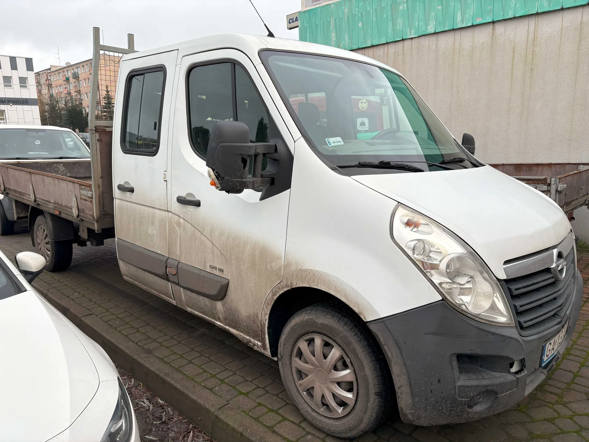 Biały, zabrudzony furgon zaparkowany na brukowanej powierzchni. W tle widoczne są inne pojazdy, w tym ciężarówka z przyczepą oraz budynki mieszkalne i przemysłowe. Na przedniej szybie furgona znajduje się naklejka. Widoczna tablica rejestracyjna.