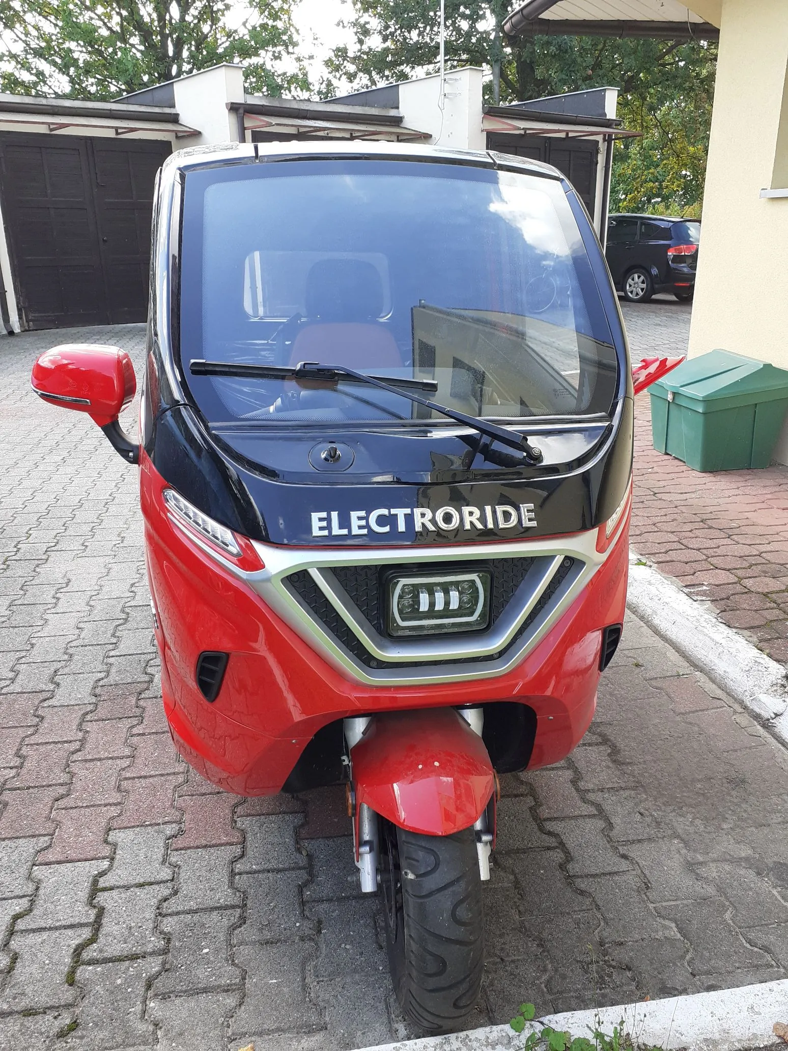 Czerwony skuter elektryczny marki Electroride zaparkowany na kostce brukowej. Pojazd ma dużą przednią szybę, czarny pas z białym napisem 'ELECTRORIDE' oraz dodatkowy reflektor LED zamontowany z przodu. W tle widoczne są garaże oraz ciemny samochód osobowy.