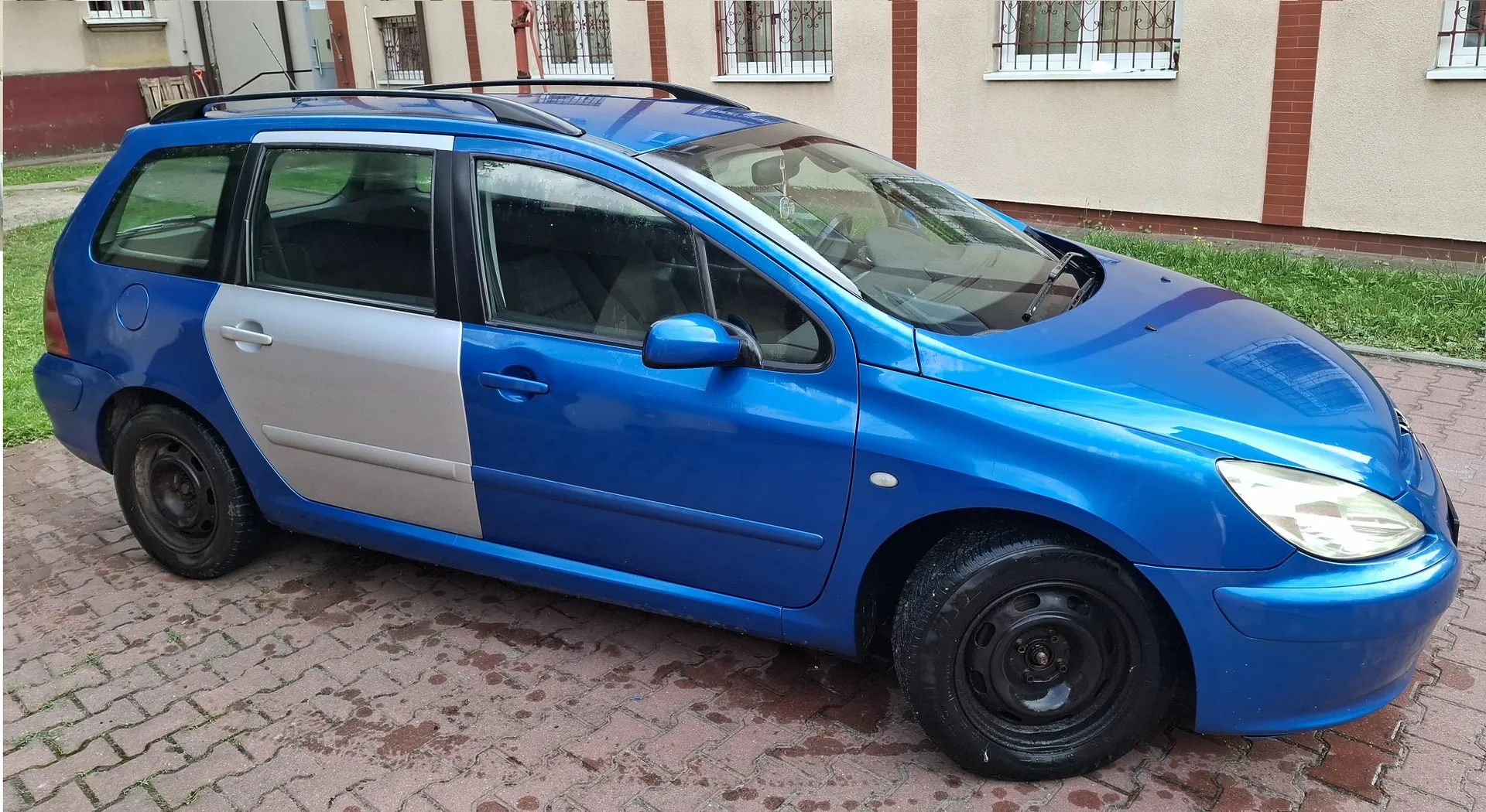 Niebieski samochód typu kombi zaparkowany na kostce brukowej przed budynkiem. Lewe tylne drzwi oraz tylny błotnik są w kolorze srebrnym, co kontrastuje z resztą niebieskiego nadwozia. Samochód posiada czarne felgi stalowe.