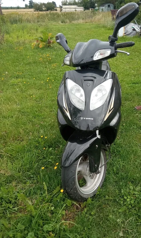 Czarny skuter marki TORQ zaparkowany na zielonej trawie z małymi żółtymi kwiatami. Widoczny jest przód pojazdu z reflektorami, lusterkami oraz przednim kołem. W tle znajduje się otwarta przestrzeń z roślinnością i budynkami w oddali.