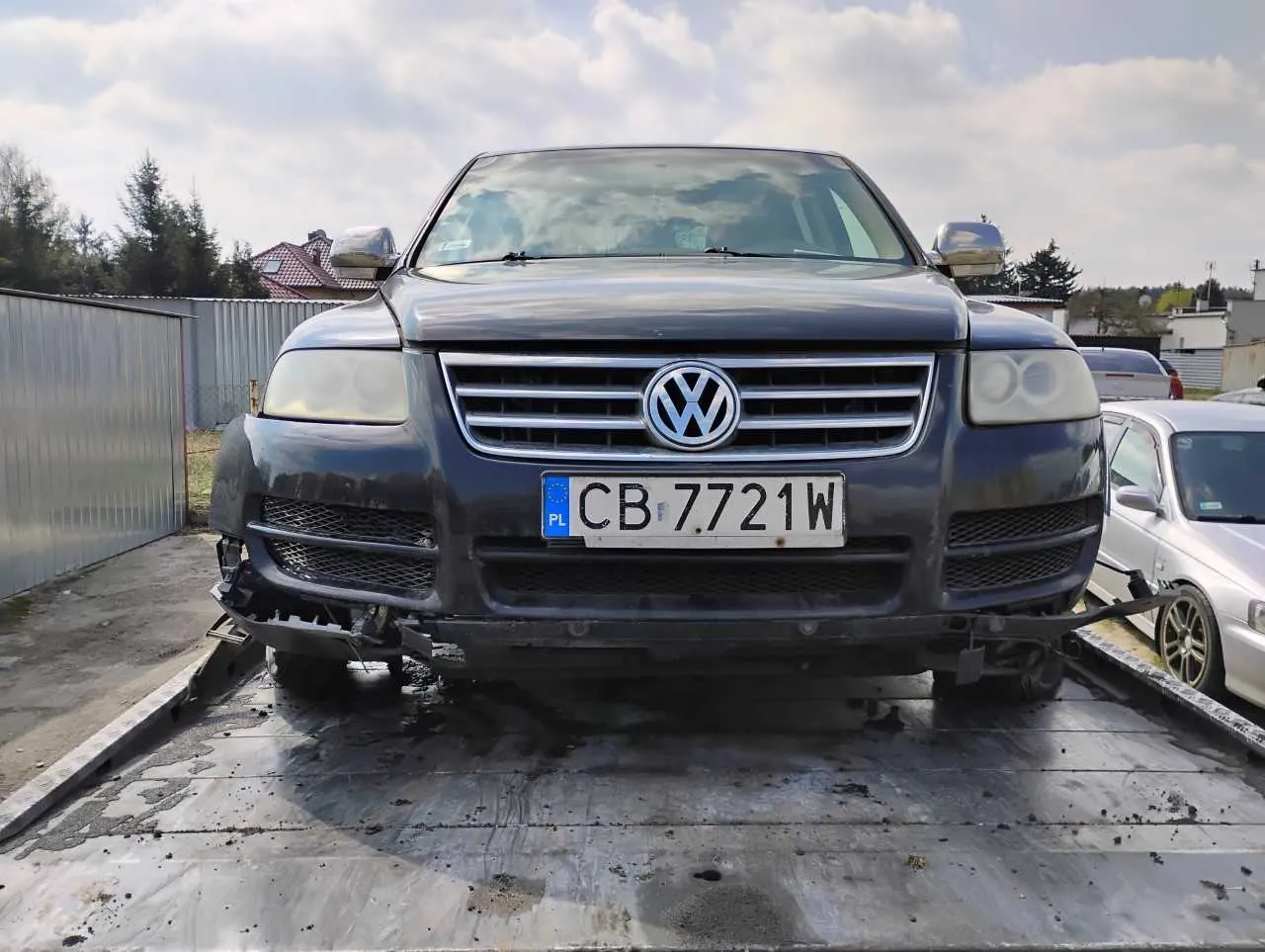 Ciemnoszary samochód marki Volkswagen (model Touareg) z uszkodzonym przodem, stojący na platformie holownika. Widoczna tablica rejestracyjna CB 7721W z niebieskim pasem PL po lewej stronie. Po prawej stronie obok znajduje się srebrny samochód. Tło: metalowe ogrodzenie, drzewa, domy z czerwonymi dachami i chmurne niebo. Podłoga platformy jest zabrudzona, a przedni zderzak Volkswagena jest częściowo uszkodzony.