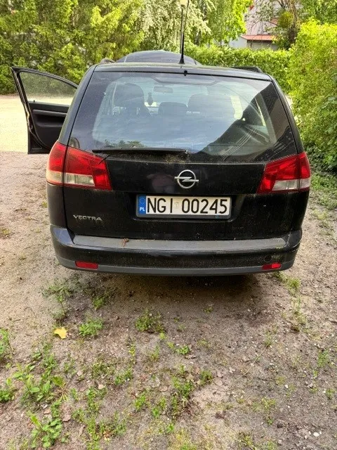Tył czarnego samochodu marki Opel Vectra zaparkowanego na piaszczystym podłożu z trawą. Samochód ma otwarte lewe tylne drzwi. Na klapie bagażnika widoczne jest logo Opela oraz polska tablica rejestracyjna o numerze NGI 00245. Po lewej stronie klapy znajduje się napis VECTRA. W tle widoczne są zielone drzewa i krzewy.