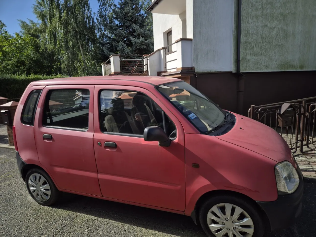 Czerwony, pięciodrzwiowy samochód osobowy zaparkowany na żwirowym podjeździe przed białym budynkiem. Auto jest ukazane z profilu od strony pasażera. W tle widoczne są zielone drzewa i fragmenty architektury budynku z balkonem.