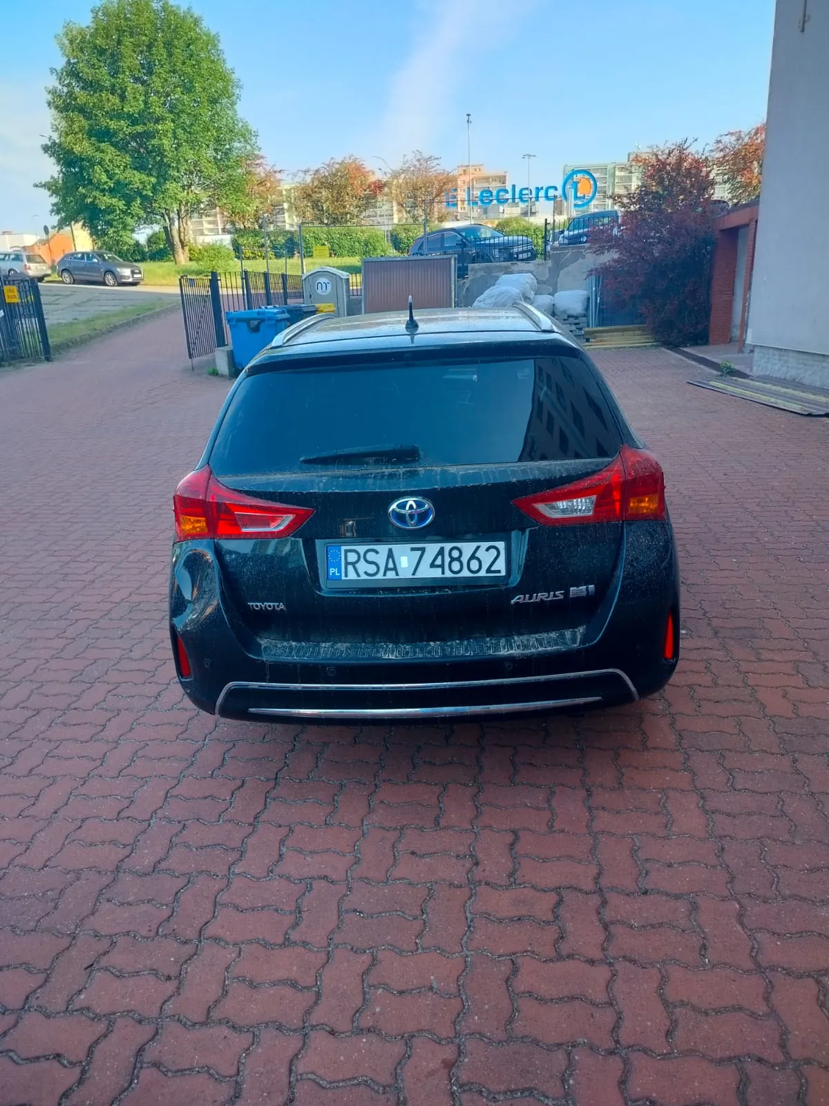 Czarny samochód Toyota Auris H1 z tablicą rejestracyjną PL RSA 74862 zaparkowany na czerwonej brukowanej powierzchni. W tle widoczny budynek z niebieskim napisem Leclerc, drzewa, inne pojazdy oraz niebo. Samochód znajduje się w centrum kadru, widok z tyłu.